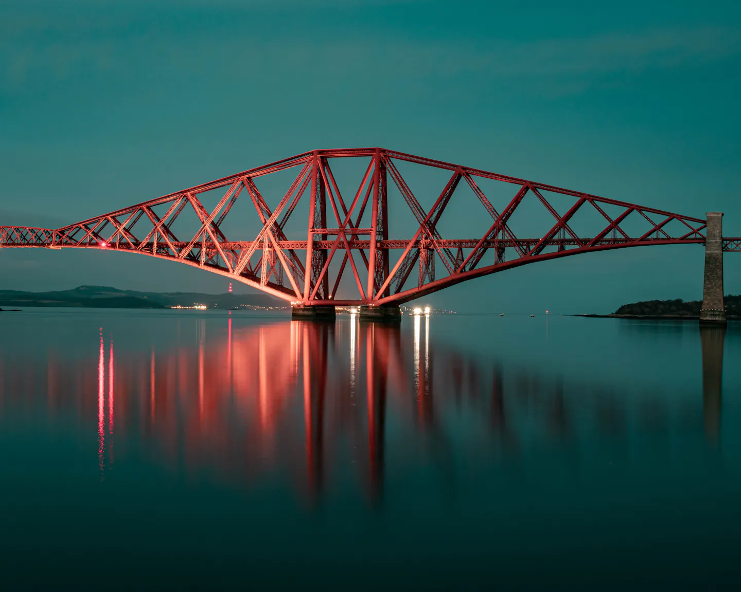 The red Forth Bridge lit at dusk, reflected in the calm Firth of Forth
