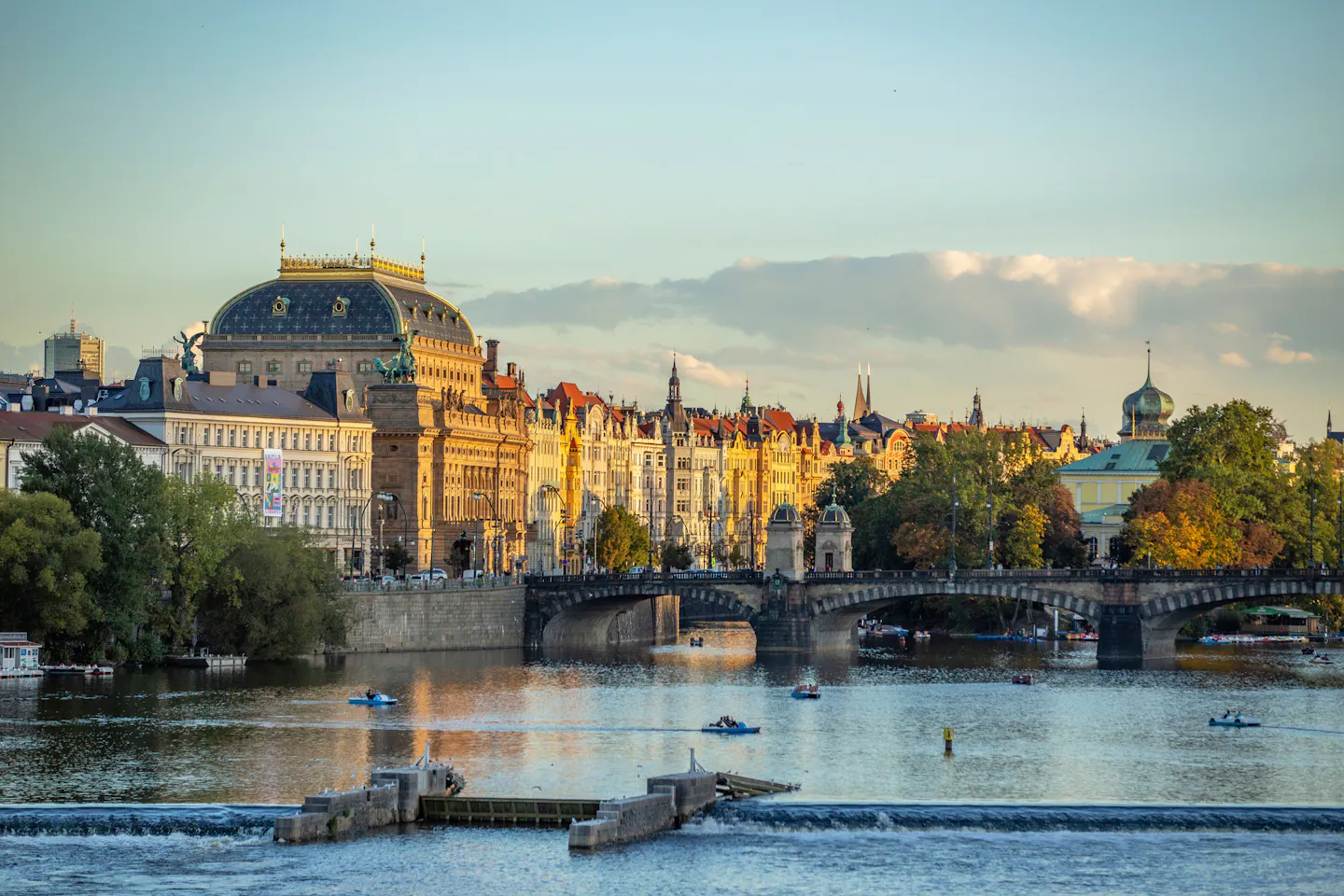 Sunlit view along the Vltava in Prague, with red-roofed historic buildings, bridges and autumn trees on the banks