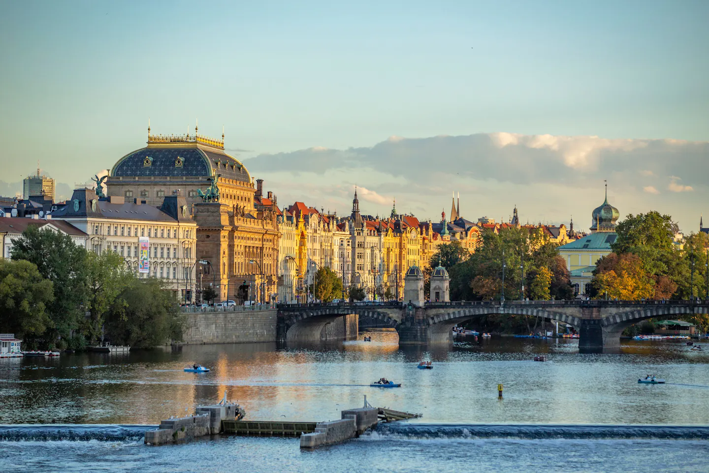 Sunlit view along the Vltava in Prague, with red-roofed historic buildings, bridges and autumn trees on the banks