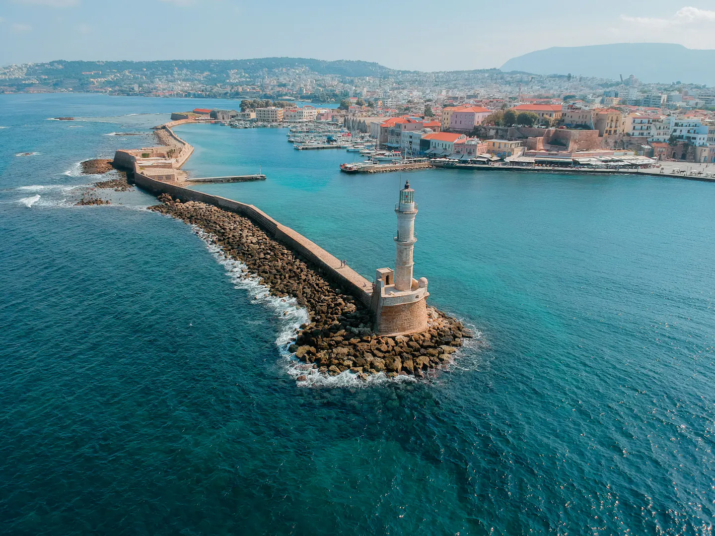 The Venetian lighthouse at the end of the stone breakwater in Chania, Crete, with the old town and turquoise water behind