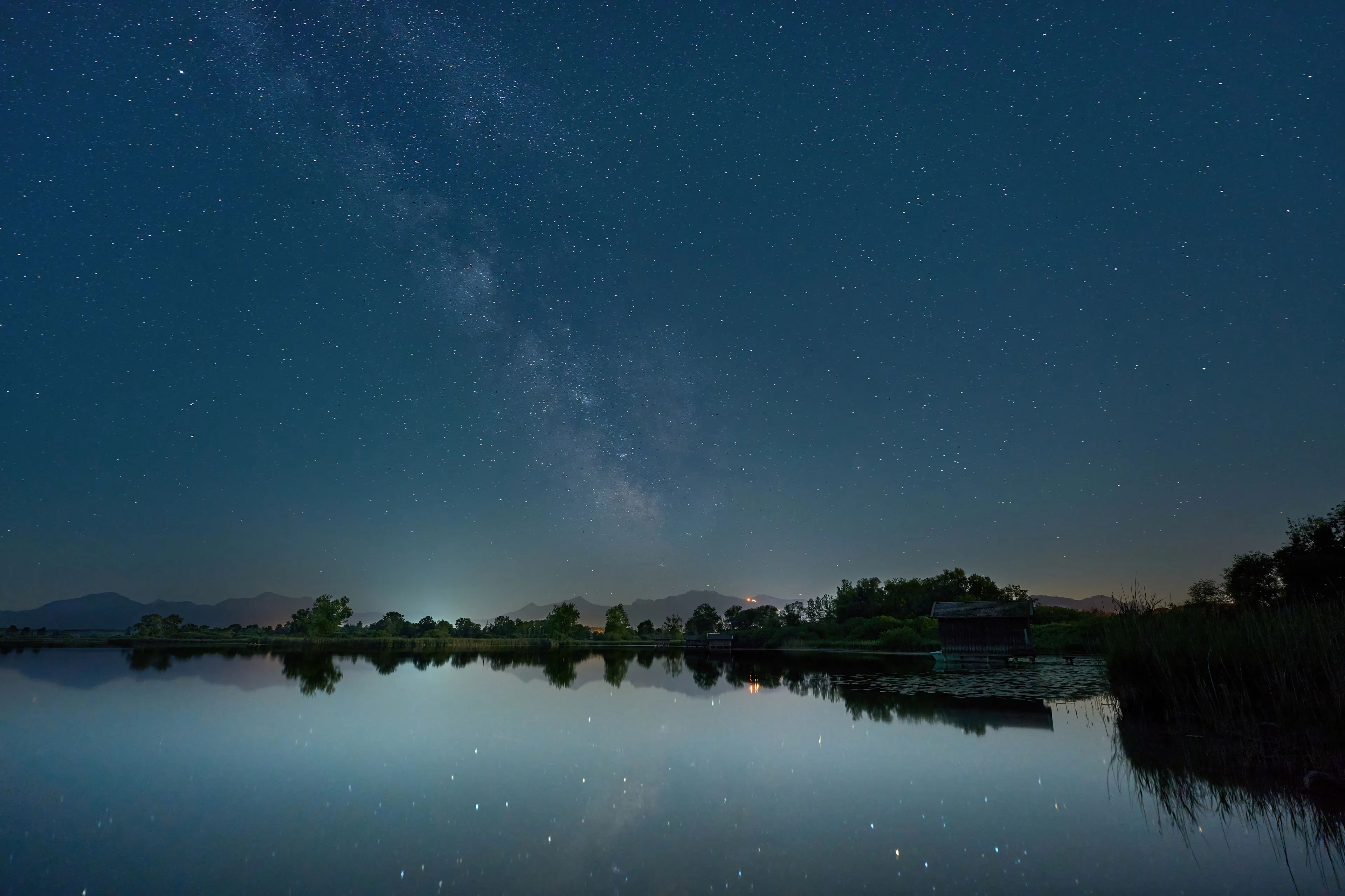 The Milky Way arching over a still lake fringed with reeds, its stars reflected in the calm water