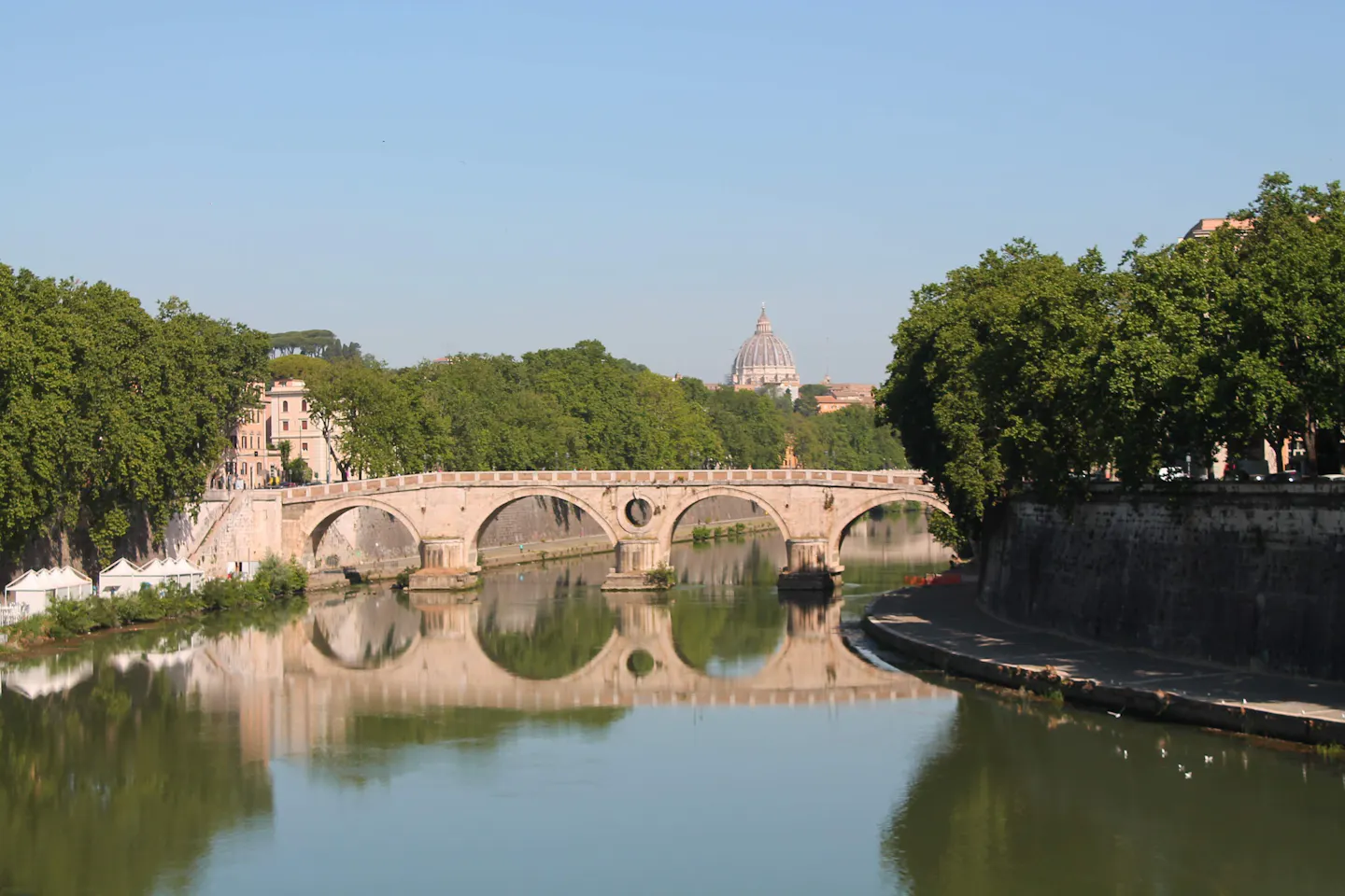 The arches of Ponte Sisto reflected in the calm Tiber on a clear morning, with the dome of St Peter's Basilica beyond the trees