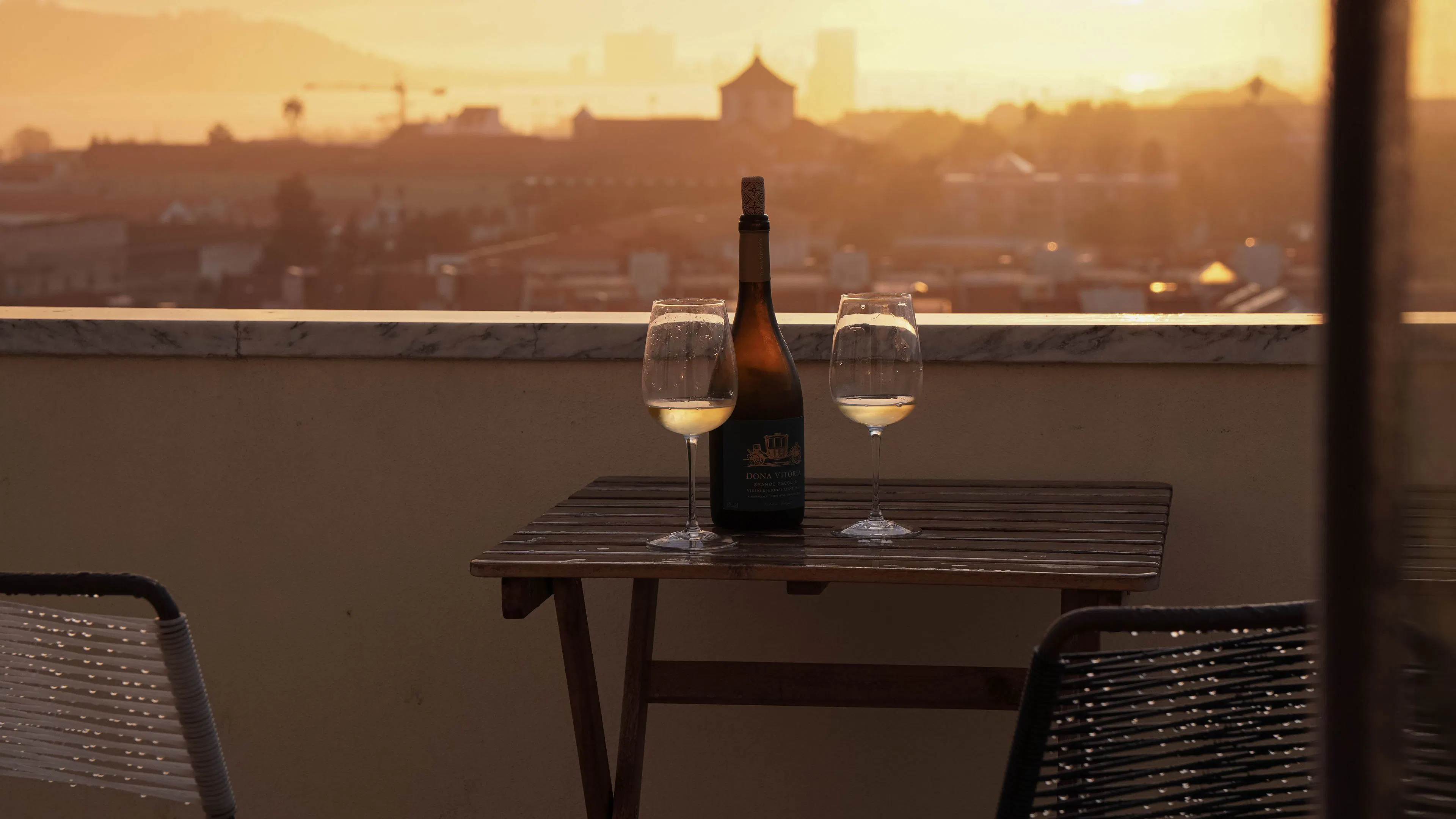 Two glasses of white wine and a bottle on a rooftop terrace table, with Lisbon's rooftops glowing in the sunset behind
