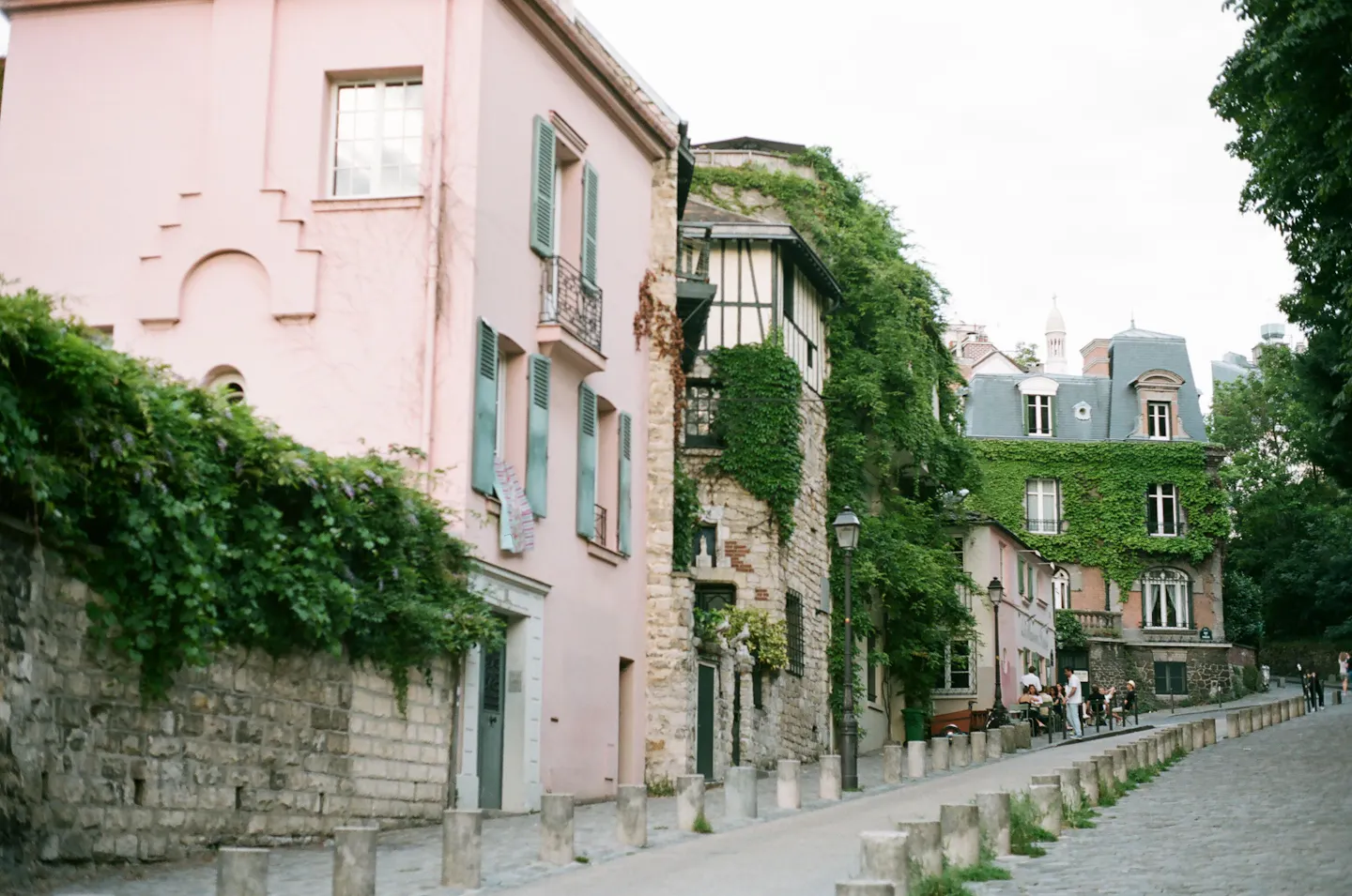A cobbled Montmartre street climbing between a large pink house with pale green shutters, an ivy-covered stone wall and creeper-clad buildings further down the hill