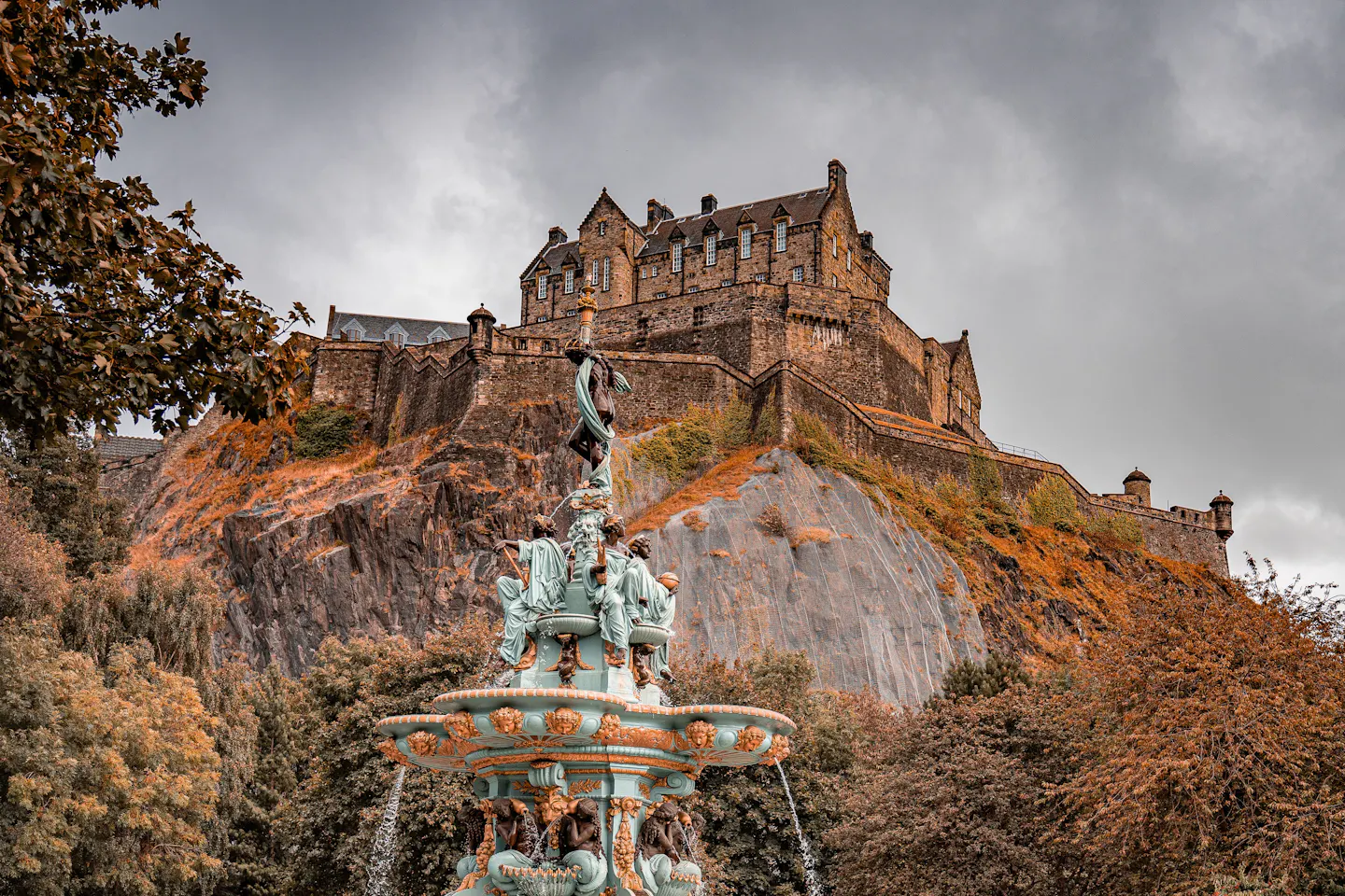 The Ross Fountain in Princes Street Gardens below Edinburgh Castle on its crag, framed by rust and gold autumn foliage under a heavy grey sky