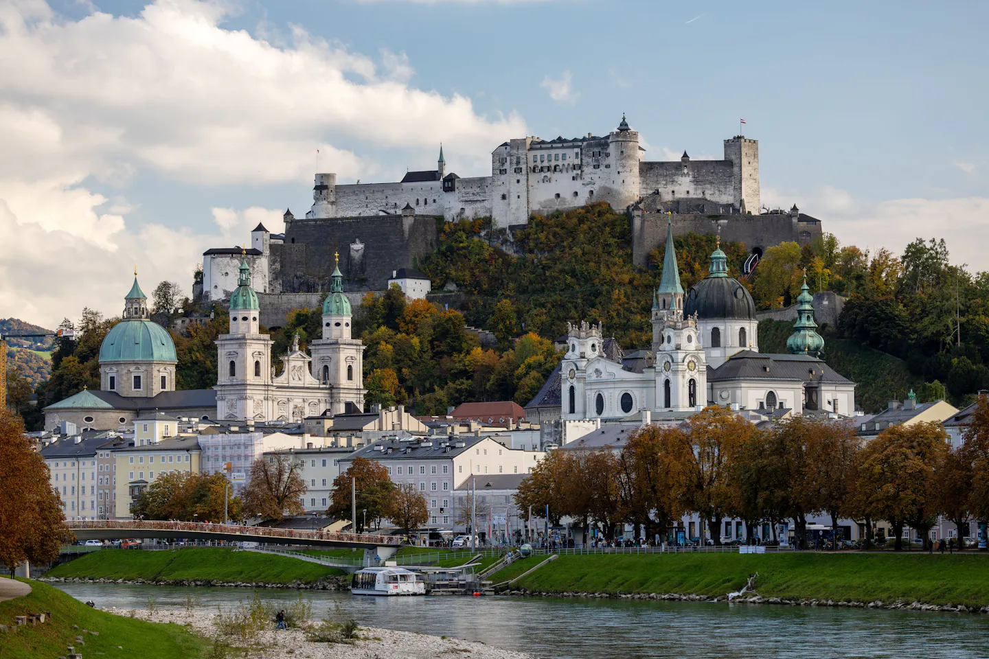 Hohensalzburg Fortress on its wooded hill above the domes and spires of Salzburg's baroque old town, with autumn trees and the Salzach in the foreground under dramatic cloud