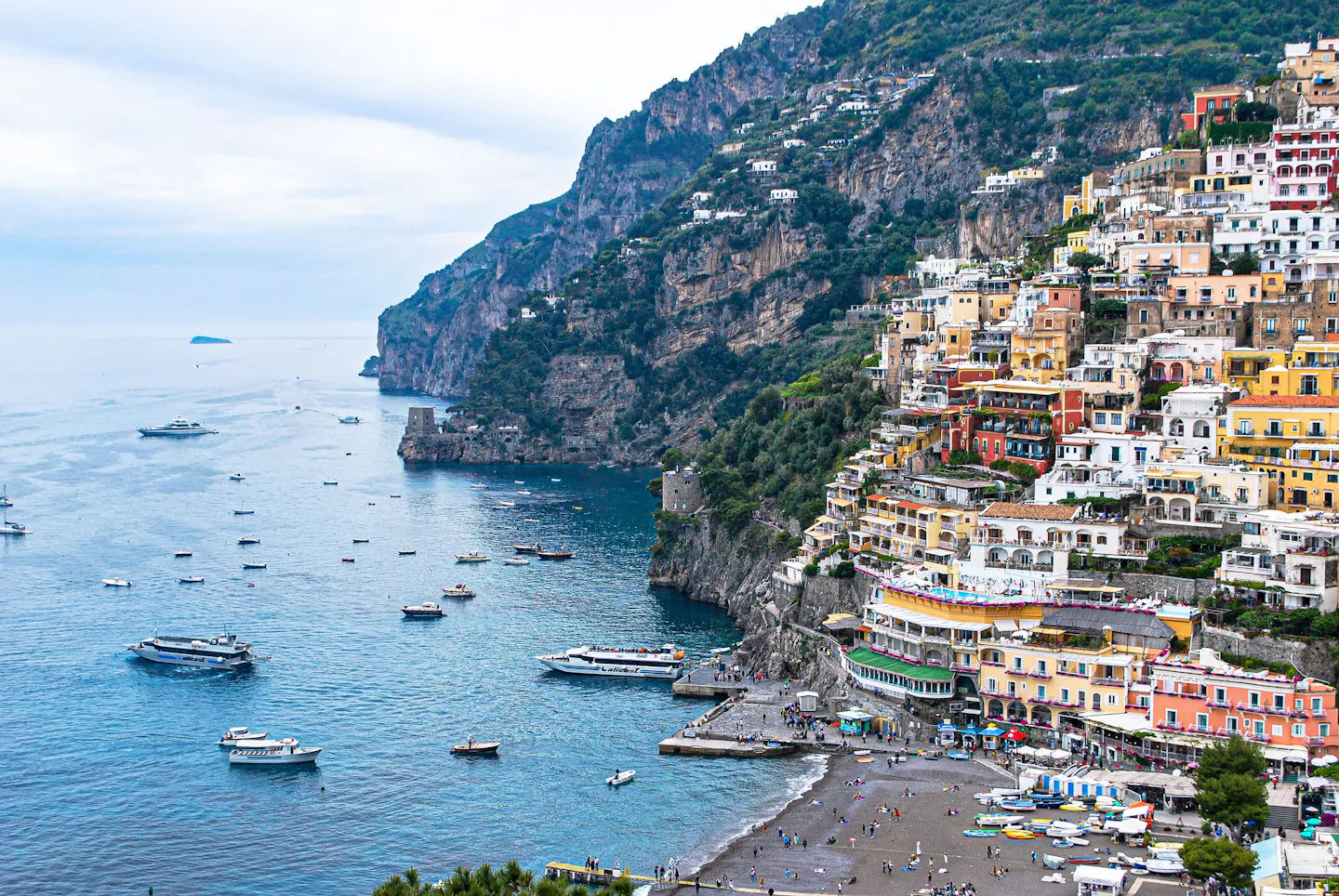 Pastel houses of Positano stacked up the cliff above the sea, seen from the water with boats moored below