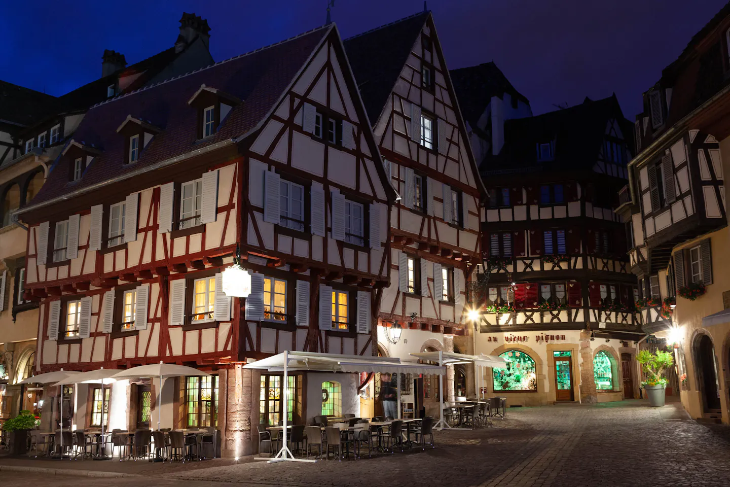 Lit half-timbered houses hung with festive garlands around an empty cobbled square in Colmar at blue hour