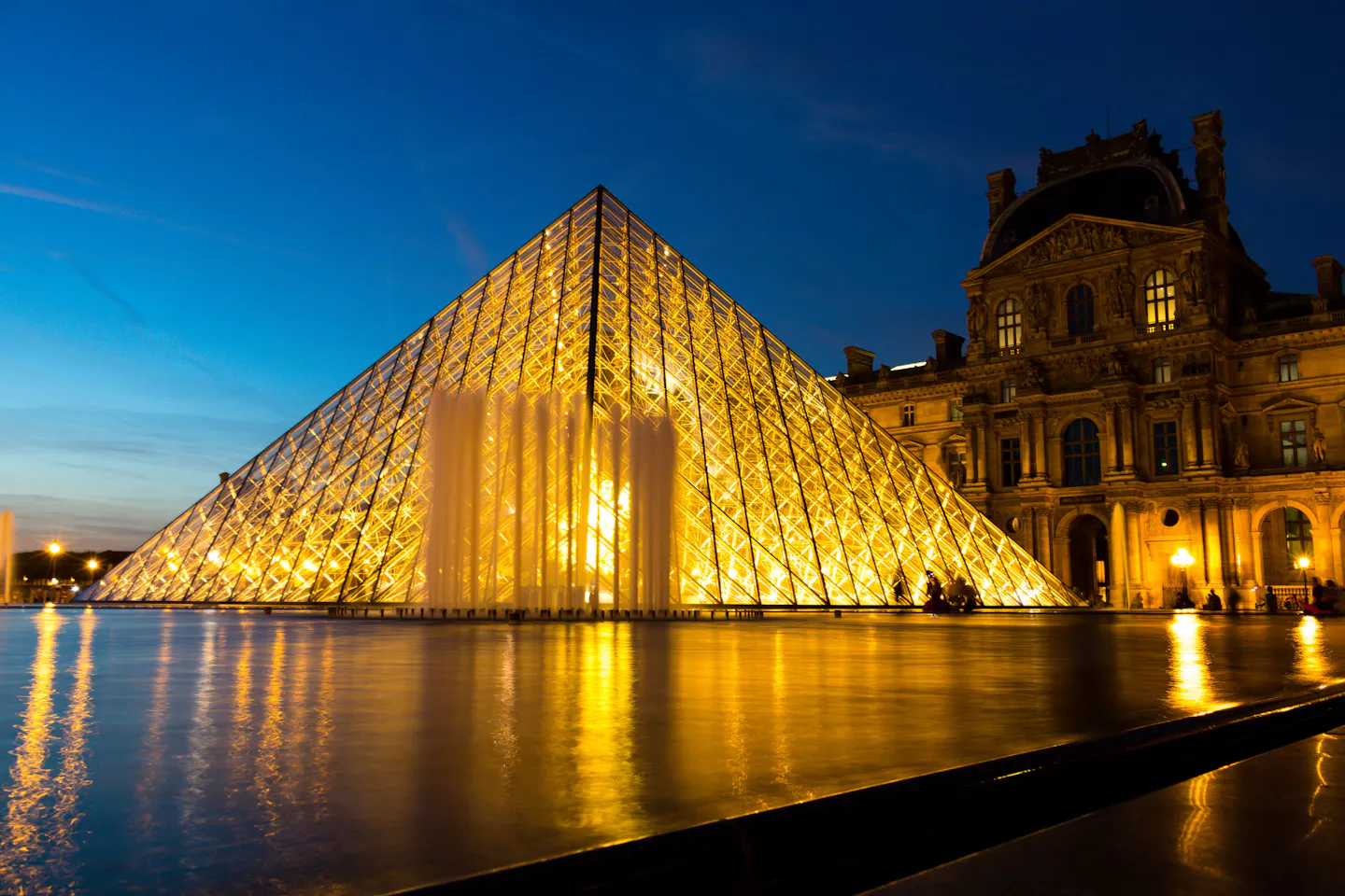 The glass pyramid of the Louvre glowing at dusk, reflected in the courtyard pool beside the palace wings