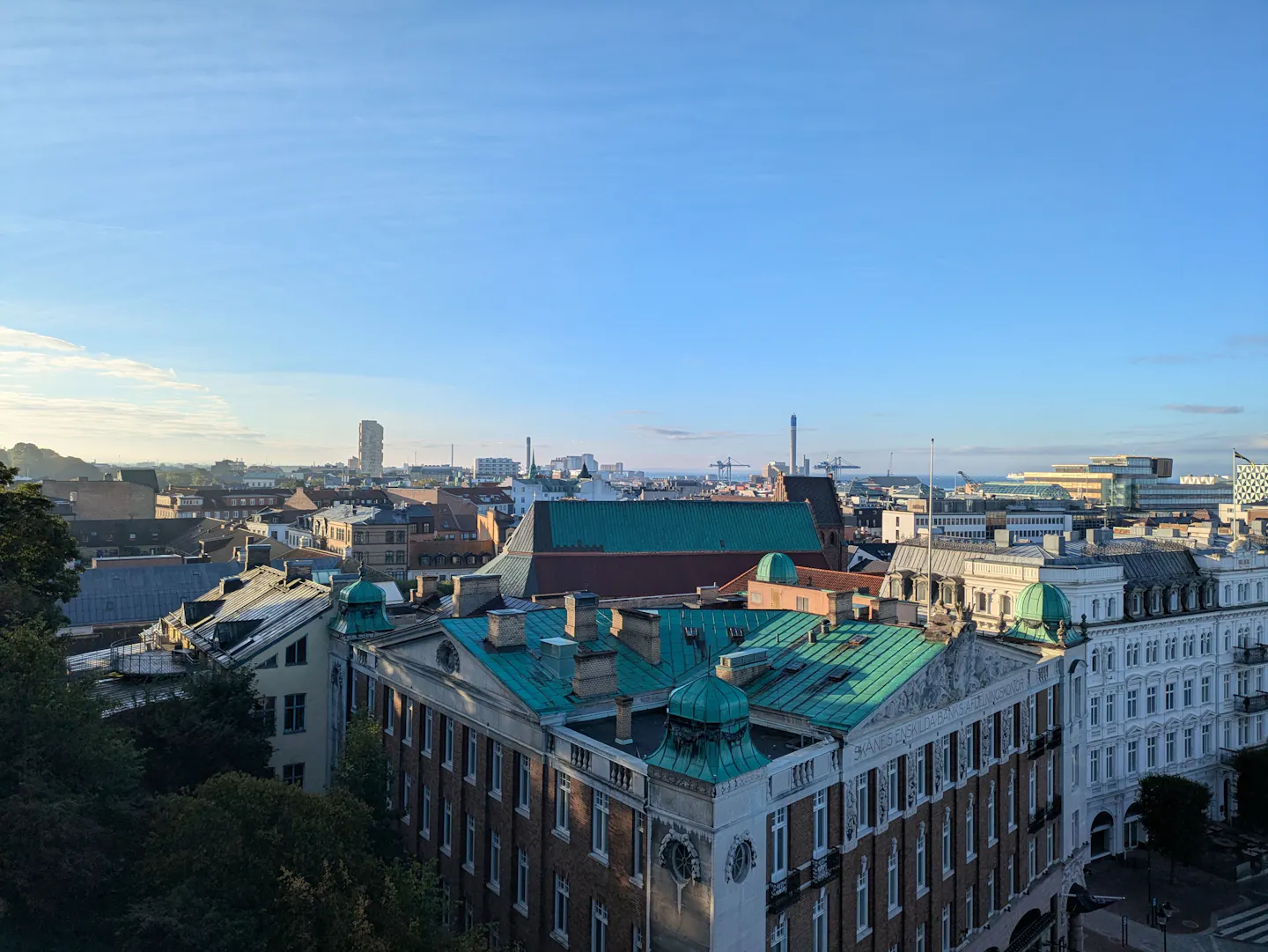 Sunlit elevated view over the historic rooftops and ornate buildings of a European city, with a modern skyline in the distance