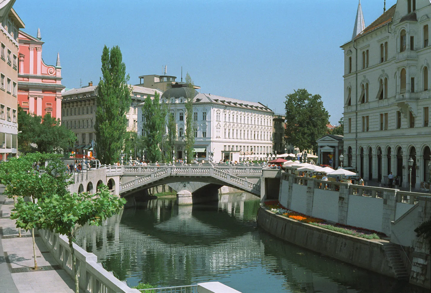 The Triple Bridge over the Ljubljanica river in Ljubljana, lined with trees and riverside cafe umbrellas
