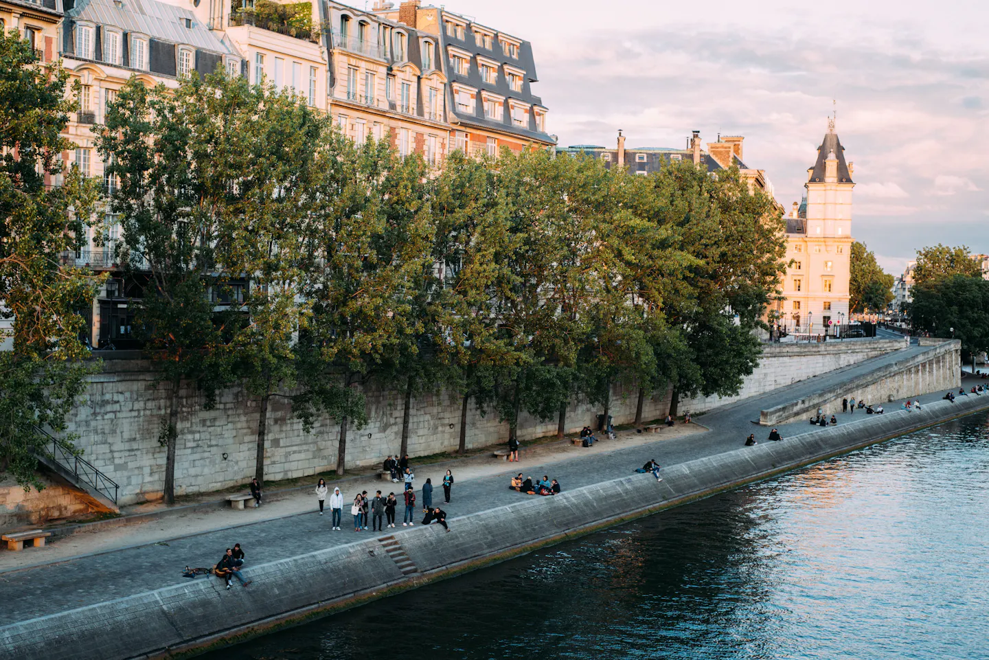 People sitting and strolling along a tree-lined Seine quay in golden evening light
