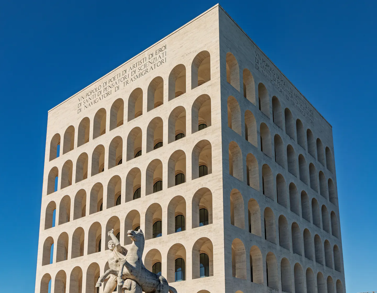 The white travertine arcaded cube of the Palazzo della Civilta Italiana in Rome's EUR district, its rows of identical arches lit by low sun against a deep blue sky, with a marble equestrian statue at its base