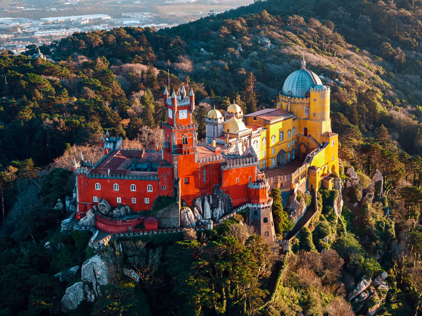 The red and yellow Pena Palace with its clock tower and grey dome standing on a wooded rocky crag above Sintra in low golden light