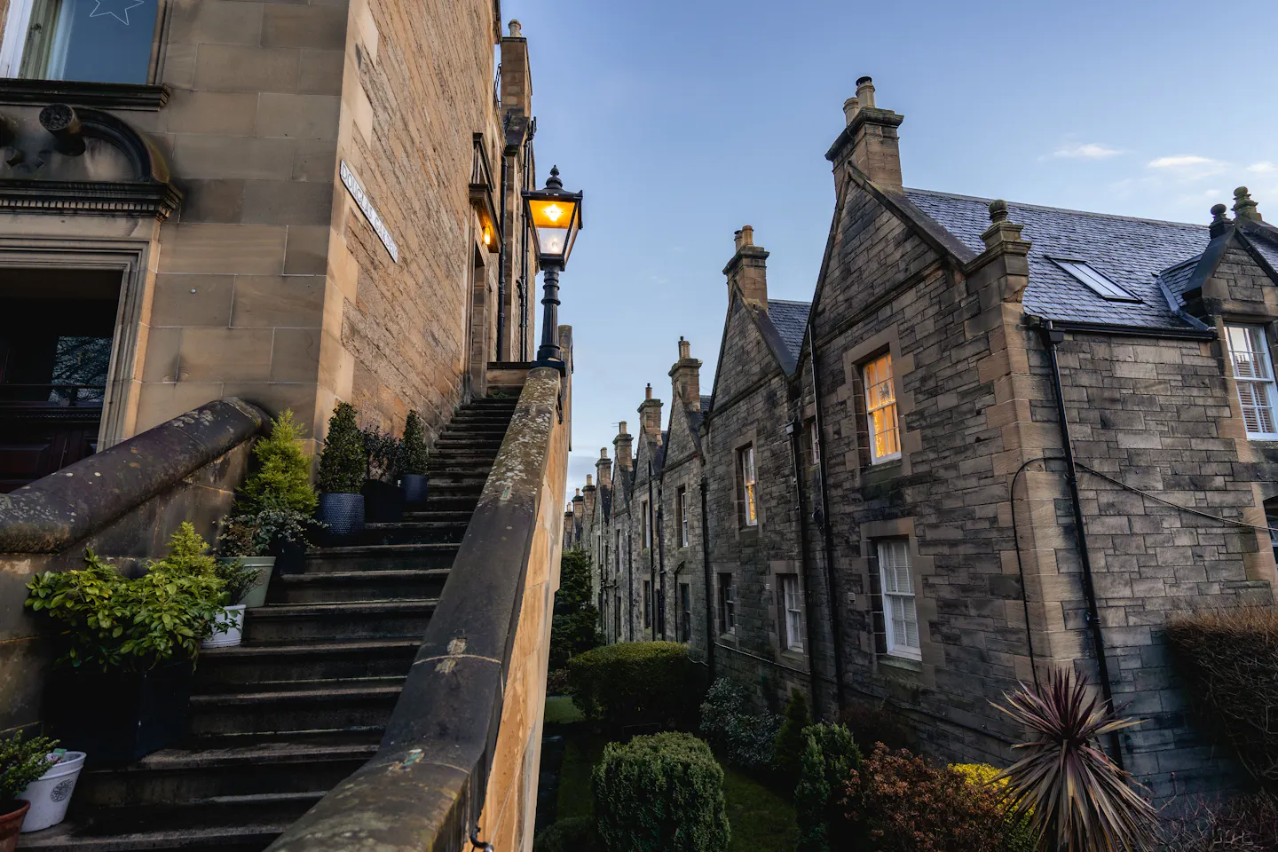 A quiet Edinburgh terrace at dusk with a lit Victorian lamp glowing against warm sandstone, stepped entrances with potted plants, and lamplight in the windows of the row beyond
