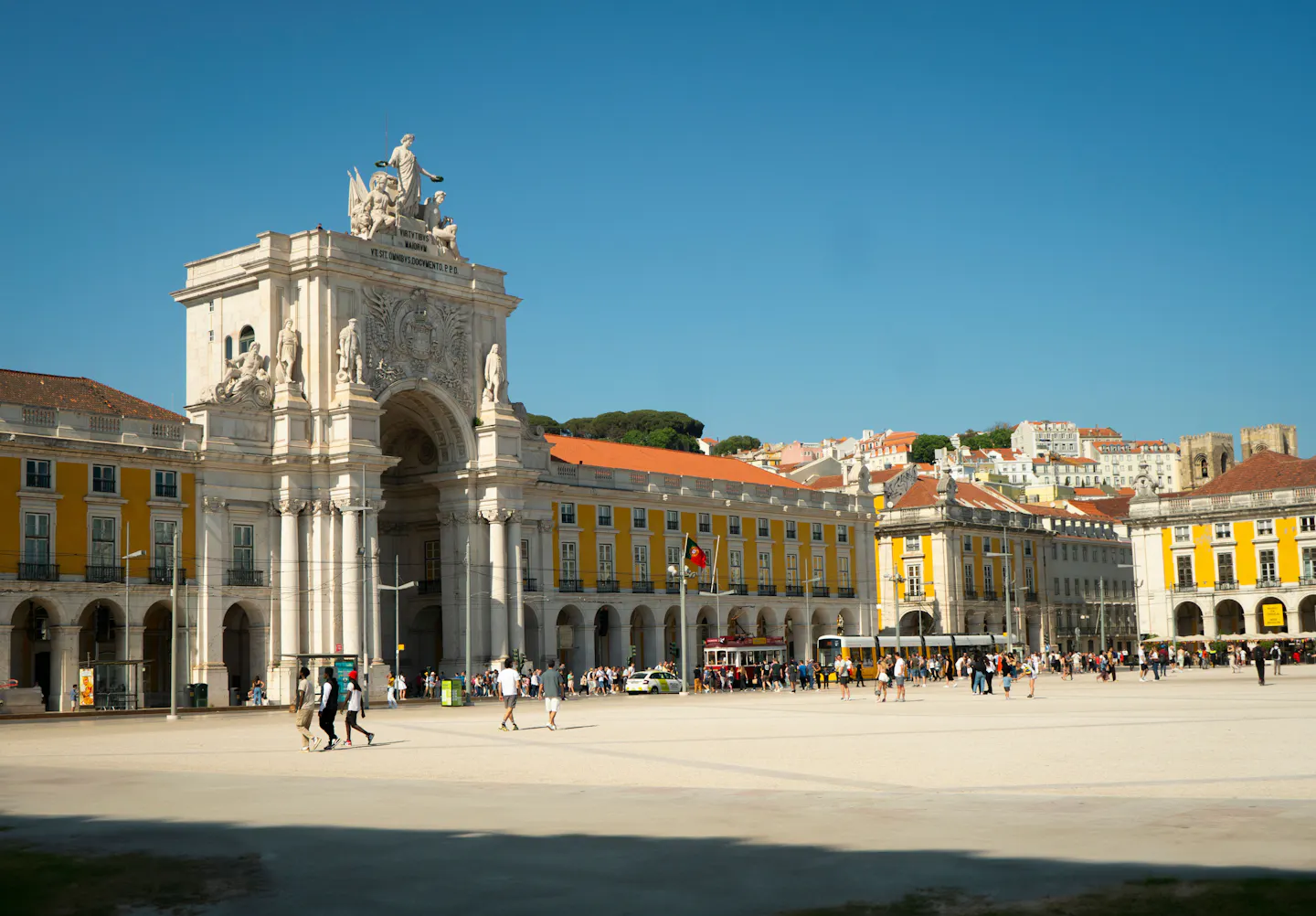 The Rua Augusta Arch and the yellow arcaded wings of Praca do Comercio under a clear blue sky, with the cathedral and castle hill rising behind