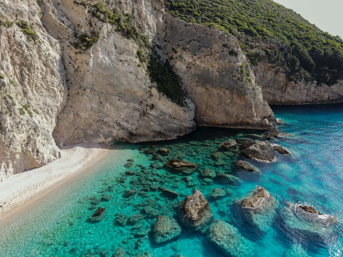 Narrow white pebble beach at the foot of a tall limestone cliff, with clear turquoise water and submerged rocks visible below the surface, seen from above