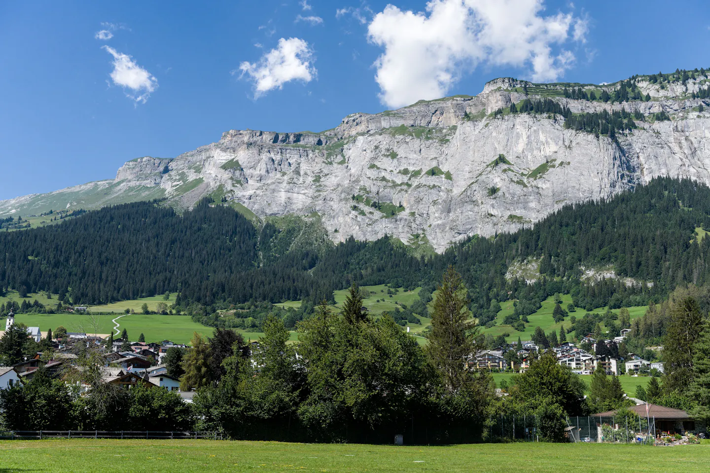 A Swiss village in green meadows below a long white limestone cliff under a bright blue sky