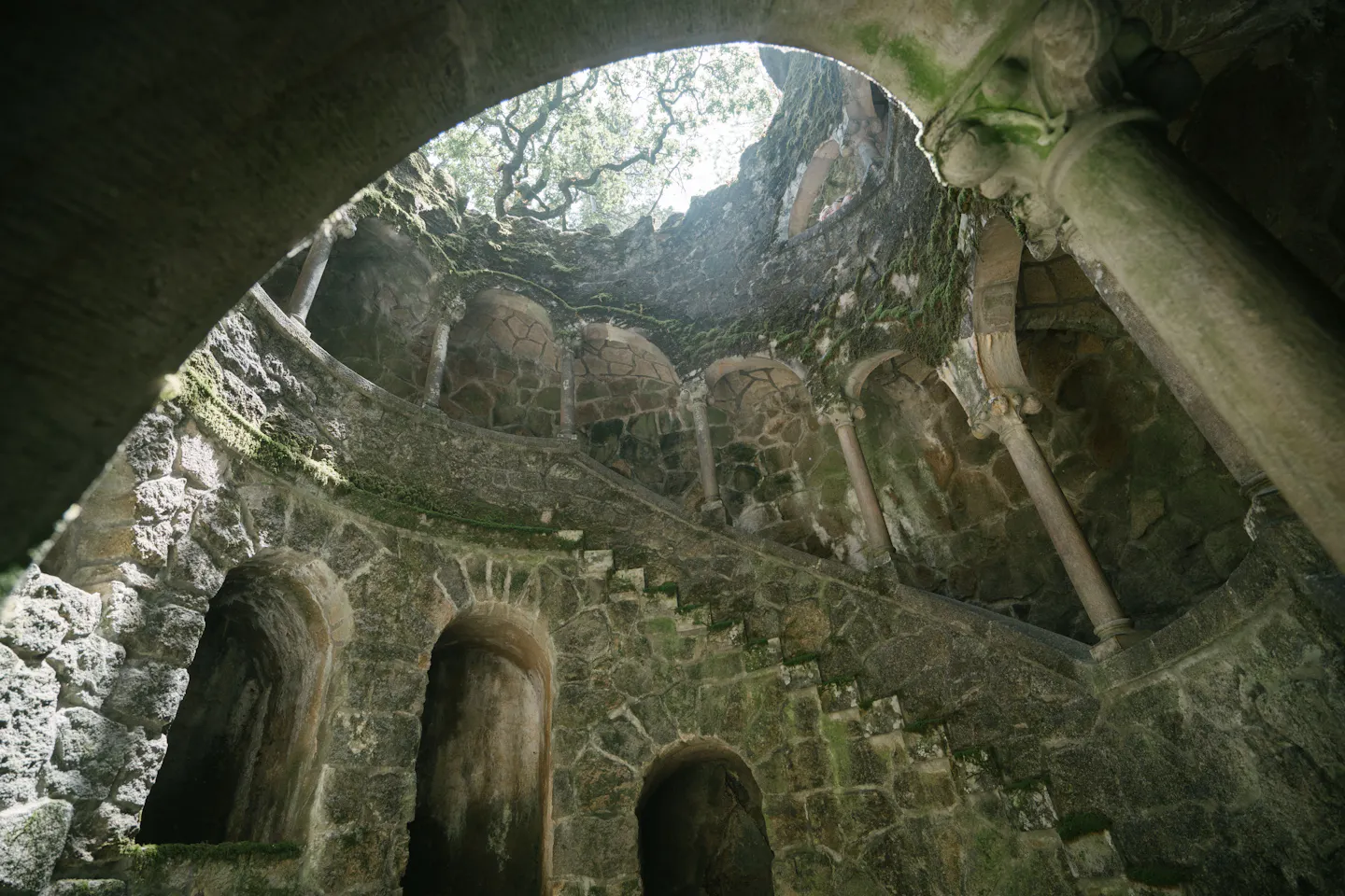 Looking up from inside the Initiation Well at Quinta da Regaleira, moss-covered stone columns and the spiral stair winding up nine levels to a ring of daylight and tree canopy