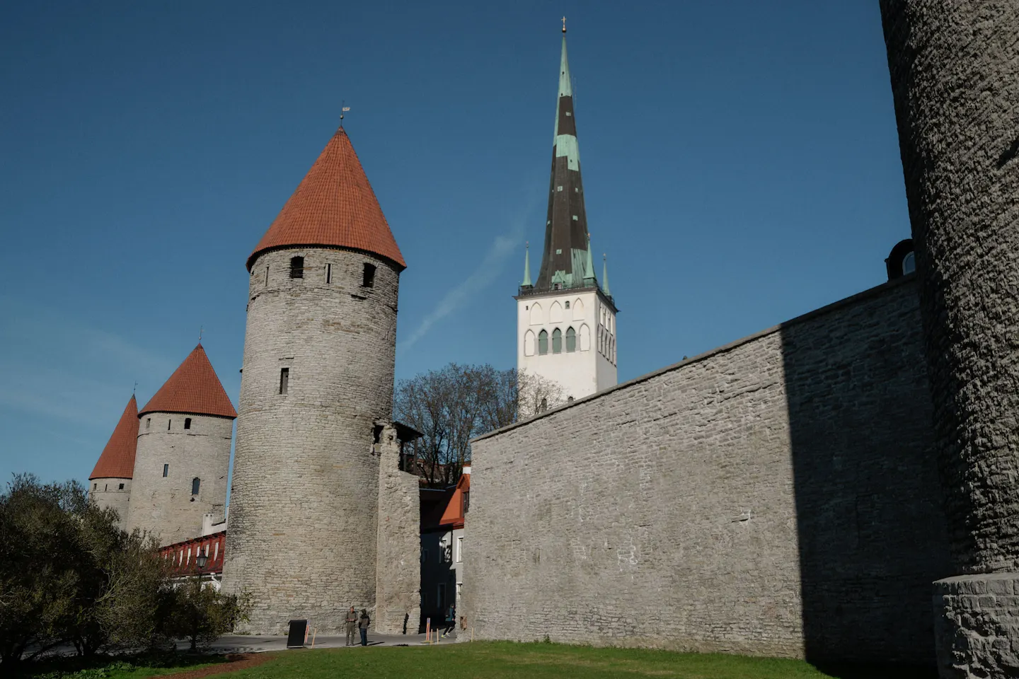 Three round red-roofed towers of Tallinn's medieval city wall stepping away beside a long sunlit limestone rampart, with the green spire of St Olaf's Church behind under a deep blue sky
