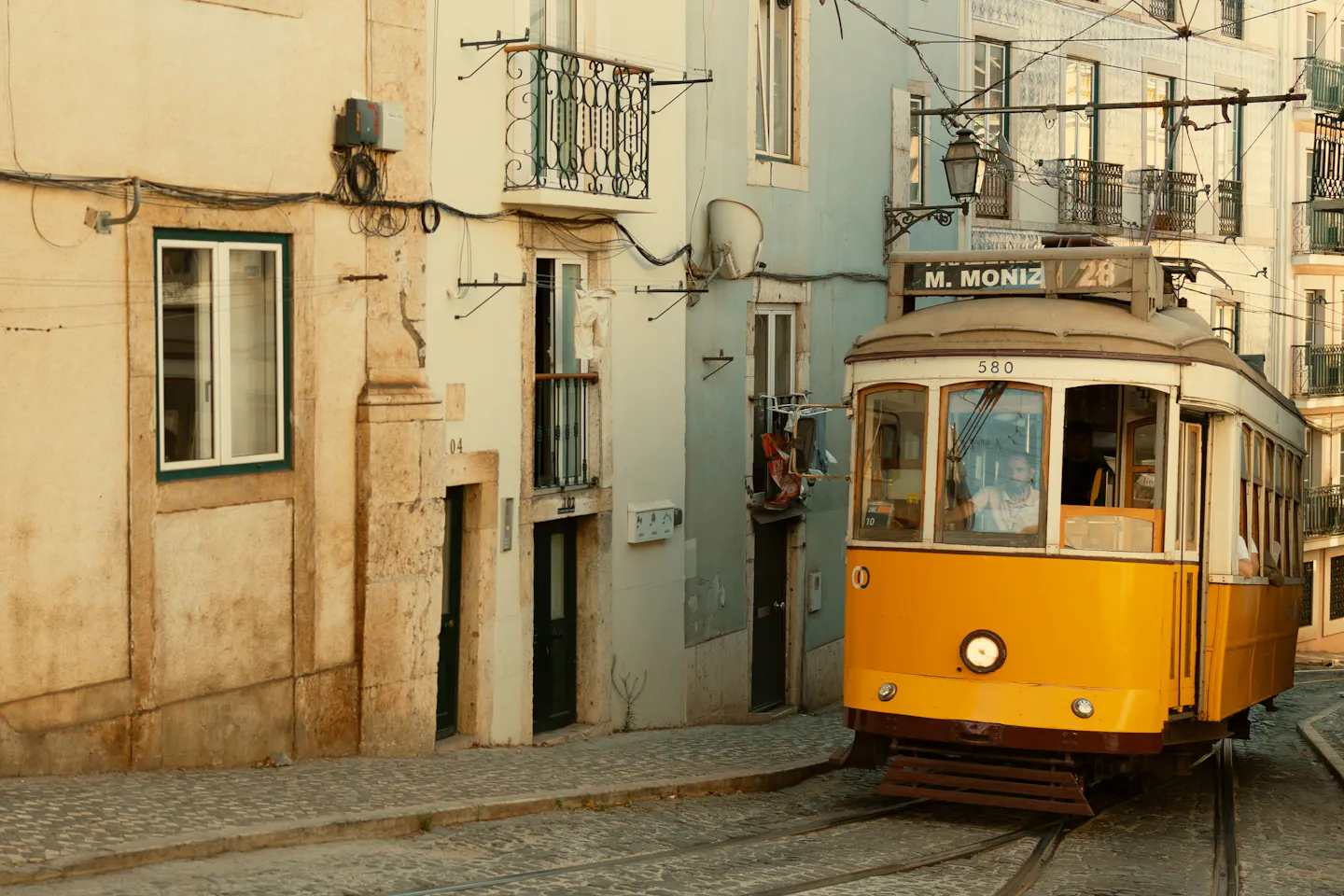 A plain yellow Lisbon tram rounding a corner on an empty cobbled street of pastel houses in warm late-afternoon light