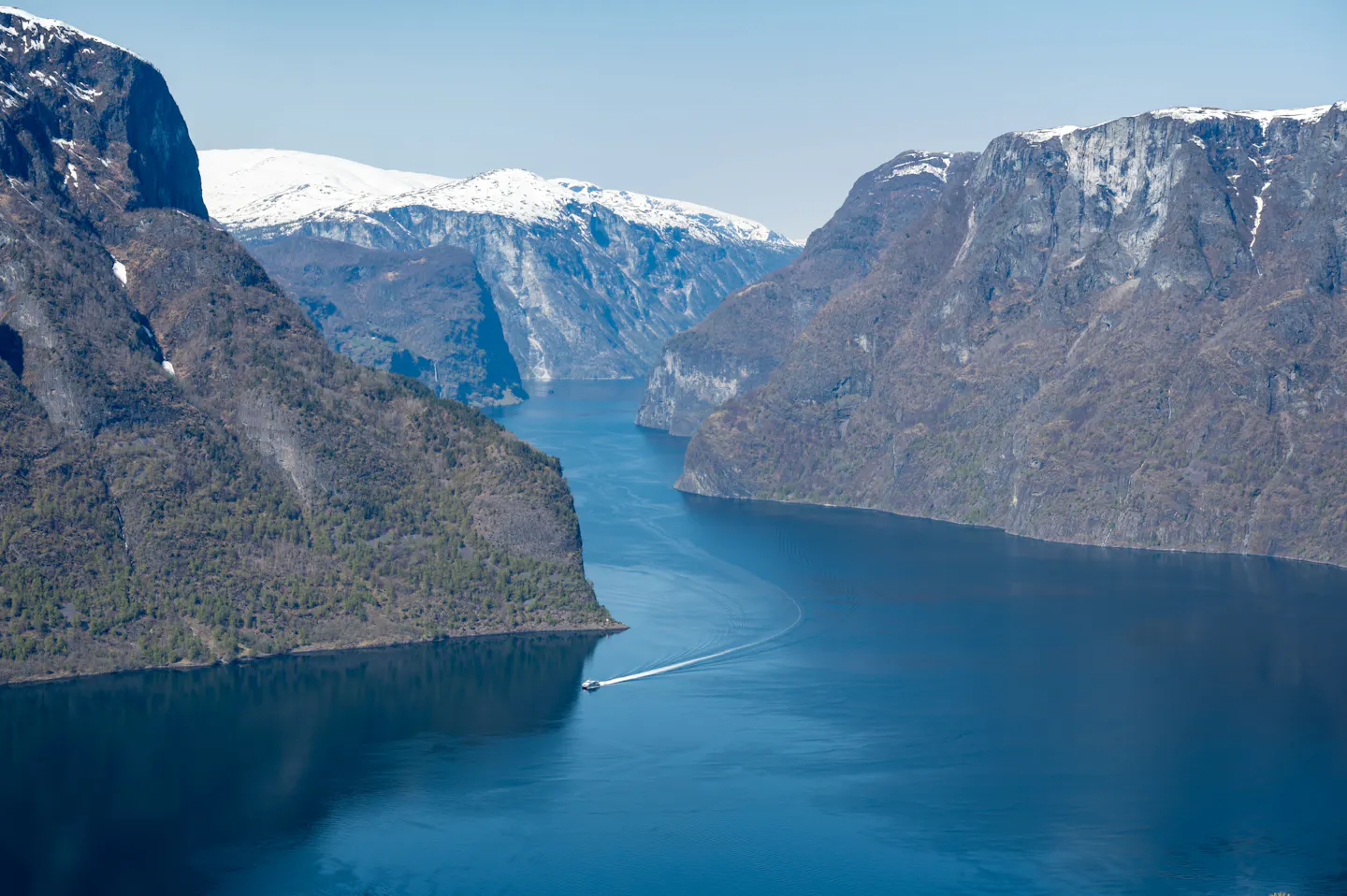 A boat leaving a wake on the deep blue Aurlandsfjord between steep snow-topped Norwegian cliffs