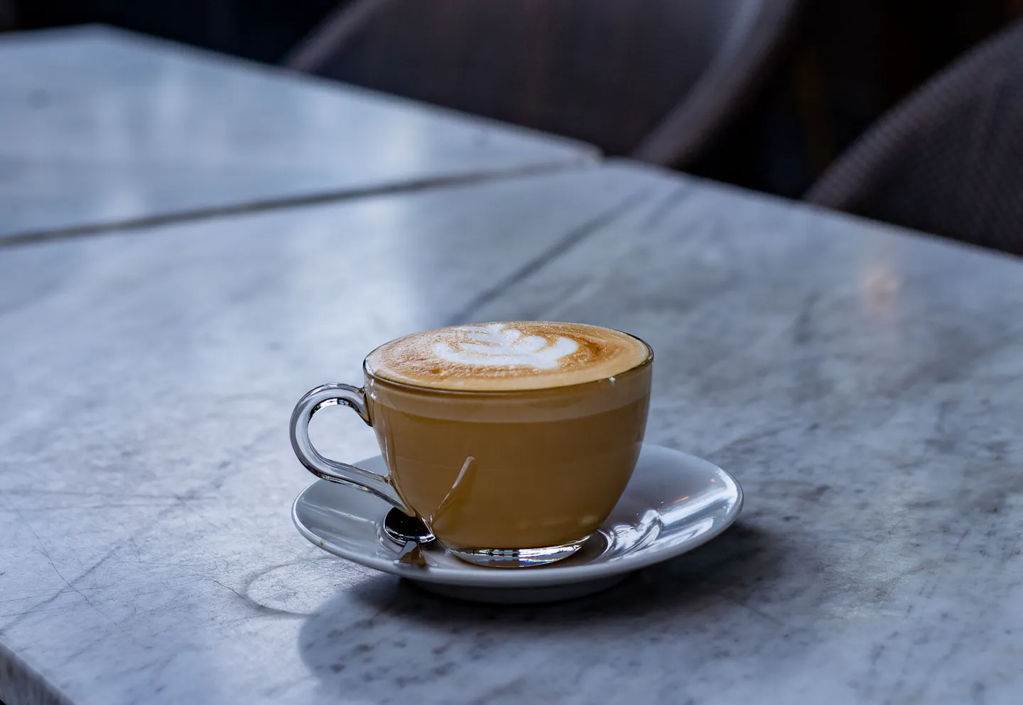 A cappuccino with leaf latte art in a white cup and saucer on a veined marble cafe table, a woven bistro chair just behind it