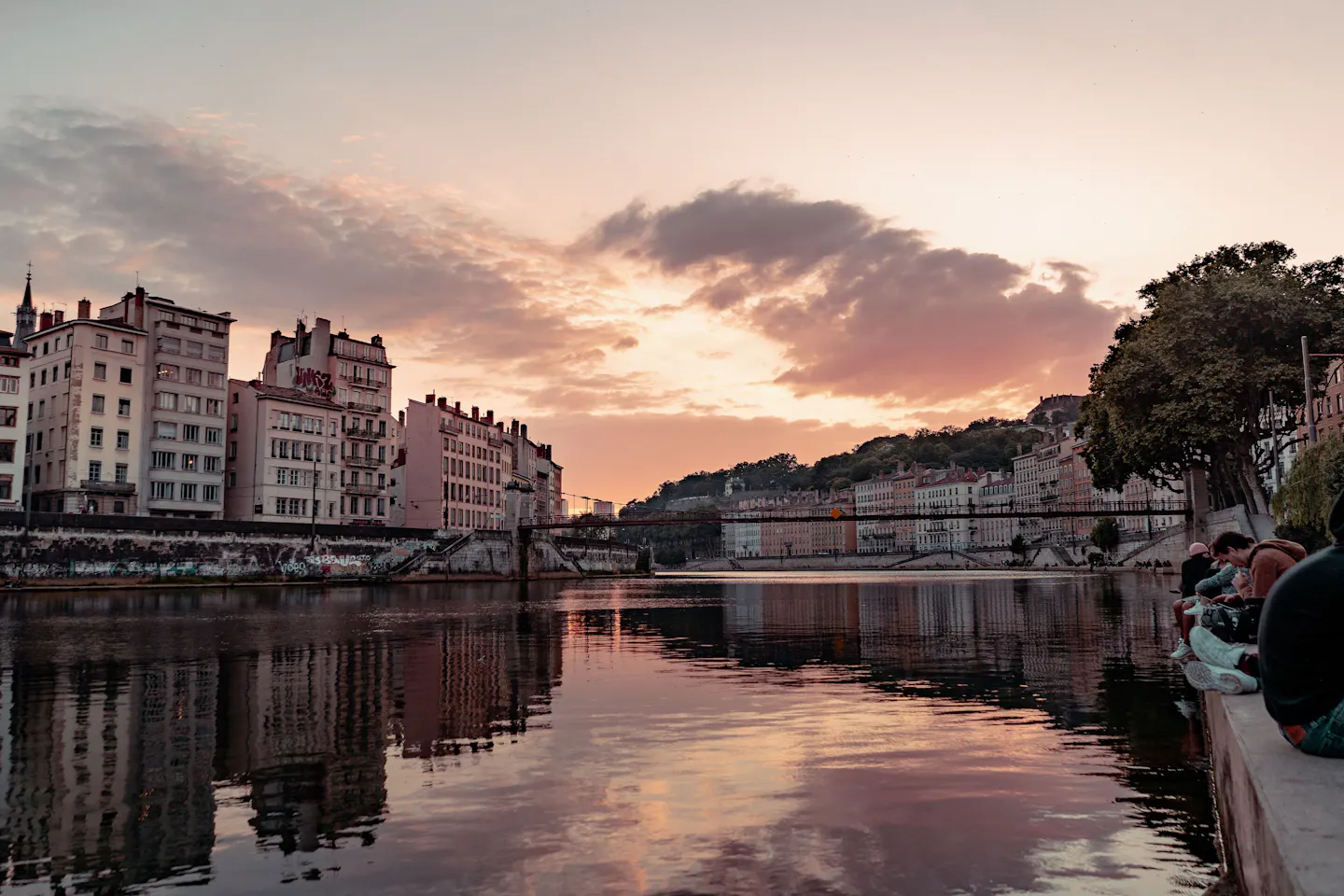 Sunset along the river in Lyon, with ornate riverside buildings, a wooded hillside and people sitting on the quay