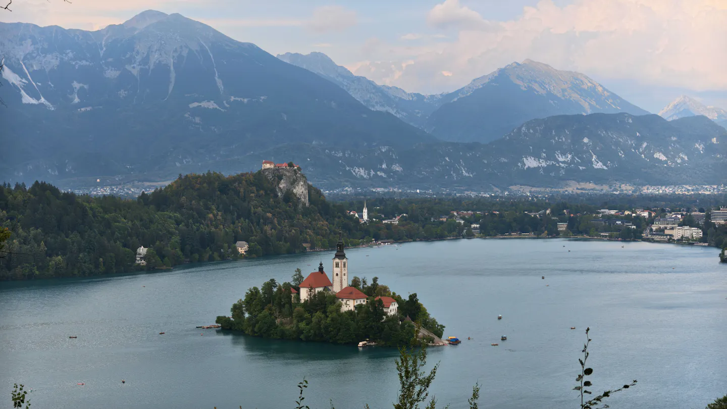 Lake Bled from a high viewpoint: the island with its church tower in the middle of the teal lake, Bled Castle on its cliff above the far shore, and the Julian Alps ranged behind