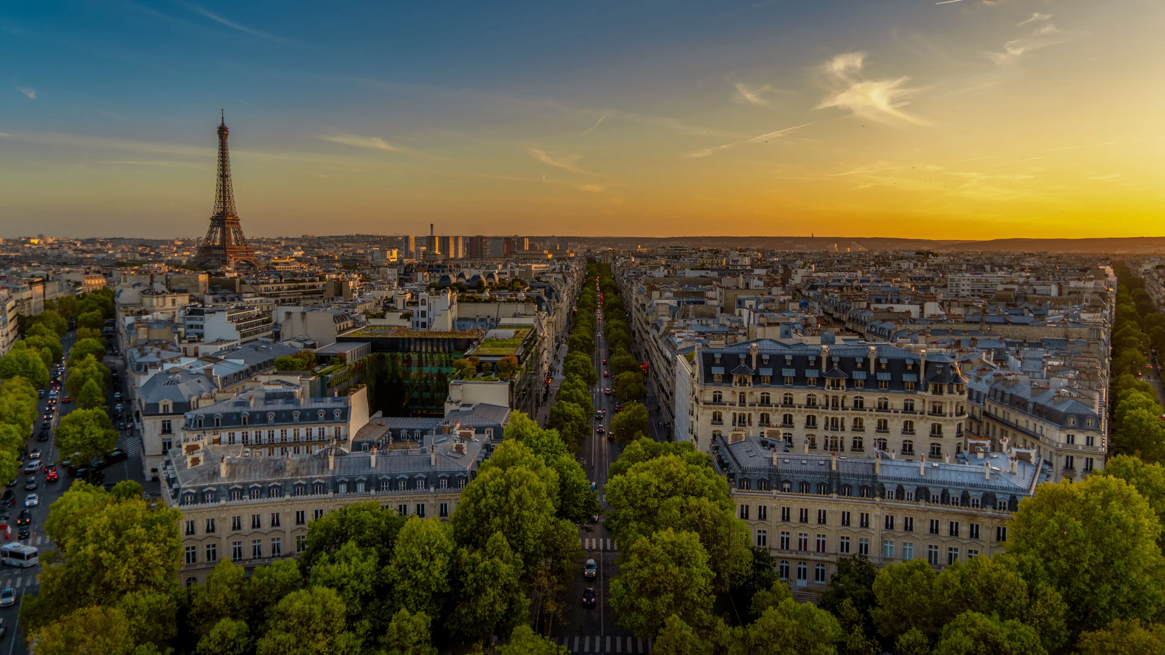Tree-lined Paris avenues and Haussmann rooftops seen from above at sunset, with the Eiffel Tower on the horizon