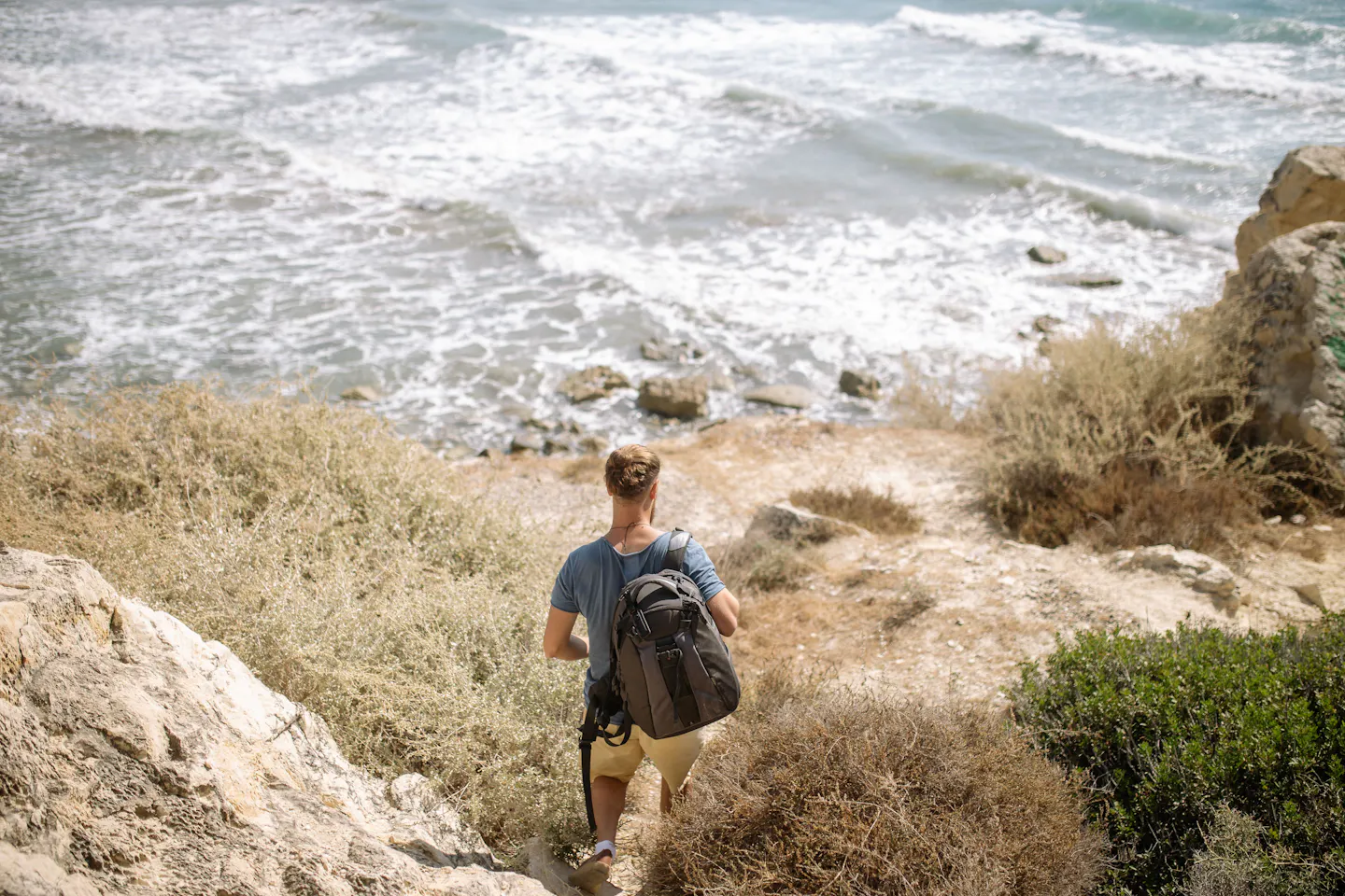 Traveler with a backpack walking down a rocky coastal path toward the sea