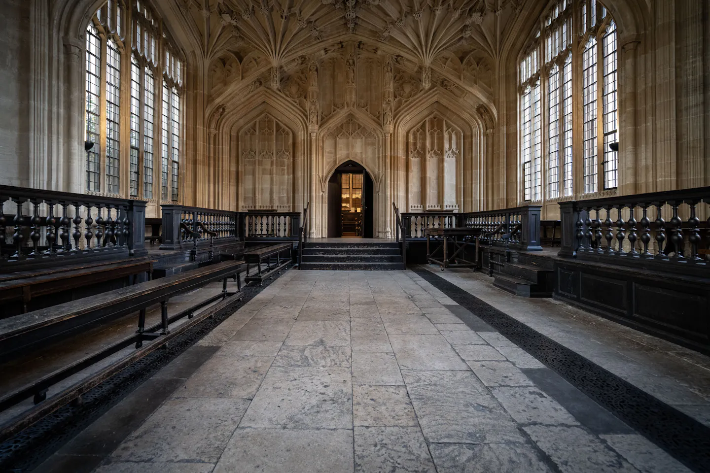 The empty fan-vaulted stone hall of Oxford's Divinity School, tall traceried windows down both sides and dark wooden benches leading to an arched doorway