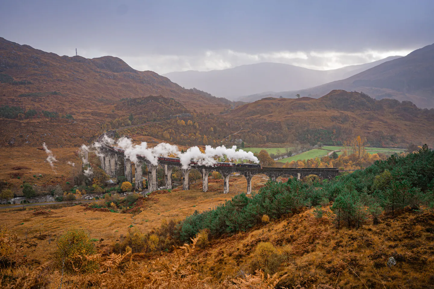 A steam train trailing smoke across the Glenfinnan Viaduct among russet Highland hills