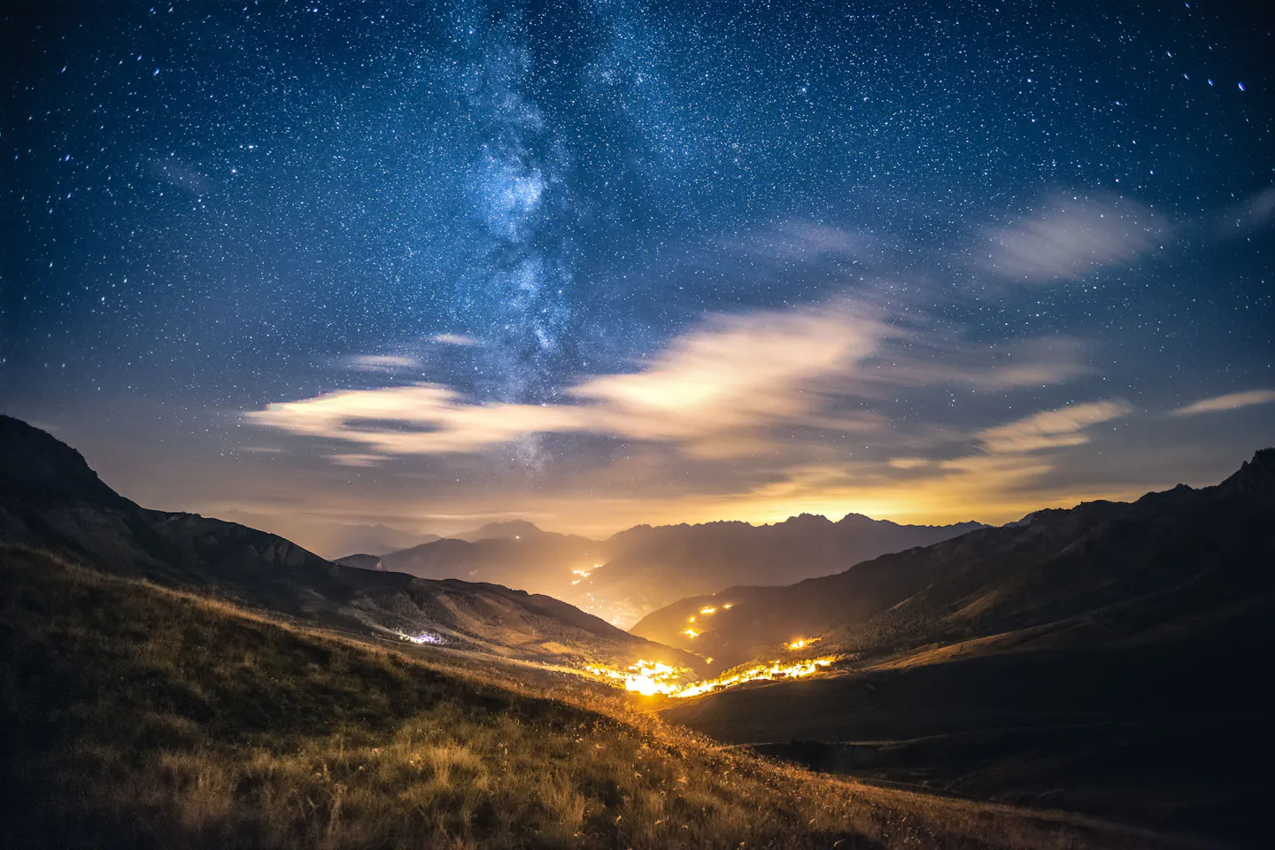 The Milky Way arching over a dark Alpine valley, with cloud bands lit gold and the warm lights of a mountain village glowing far below