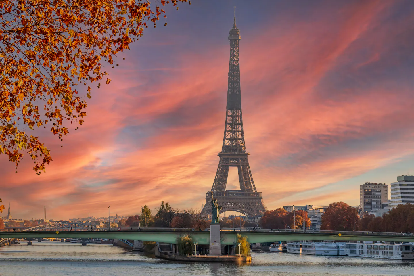 The Eiffel Tower above the Seine at sunset, framed by autumn leaves under a pink and orange sky