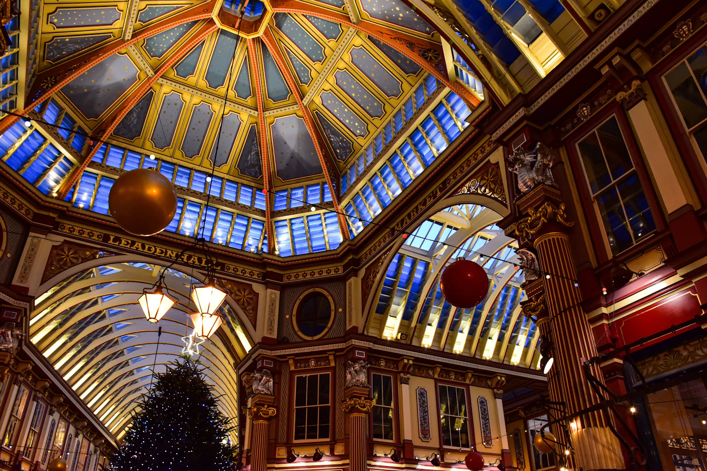 The painted blue and gold dome of Leadenhall Market at night, gold stars across the ceiling panels, hanging lanterns and red baubles, with a lit Christmas tree below