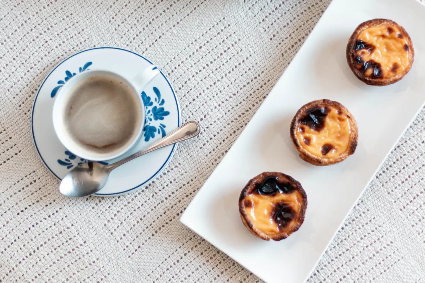 Three pasteis de nata on a white tray beside a cup of coffee on a blue-and-white saucer, seen from above