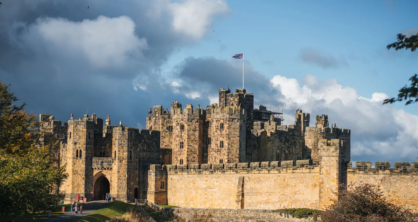 Alnwick Castle's keep and curtain walls glowing in low autumn sunlight beneath a dark storm sky, a Union flag flying from the tower and parkland grass sloping away in front
