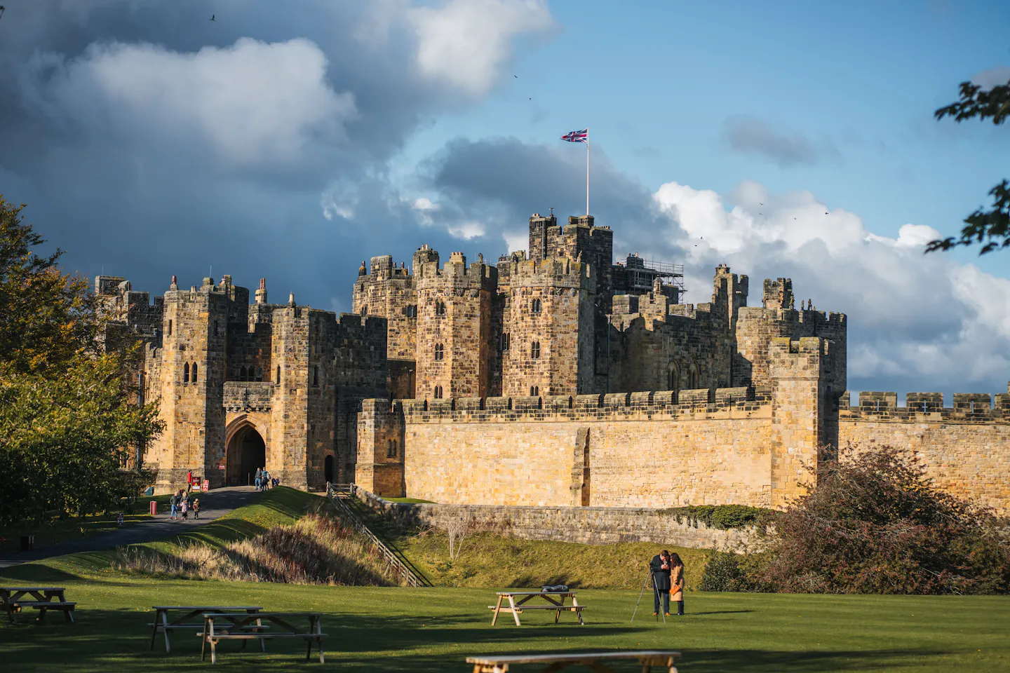 Alnwick Castle's keep and curtain walls glowing in low autumn sunlight beneath a dark storm sky, a Union flag flying from the tower and parkland grass sloping away in front