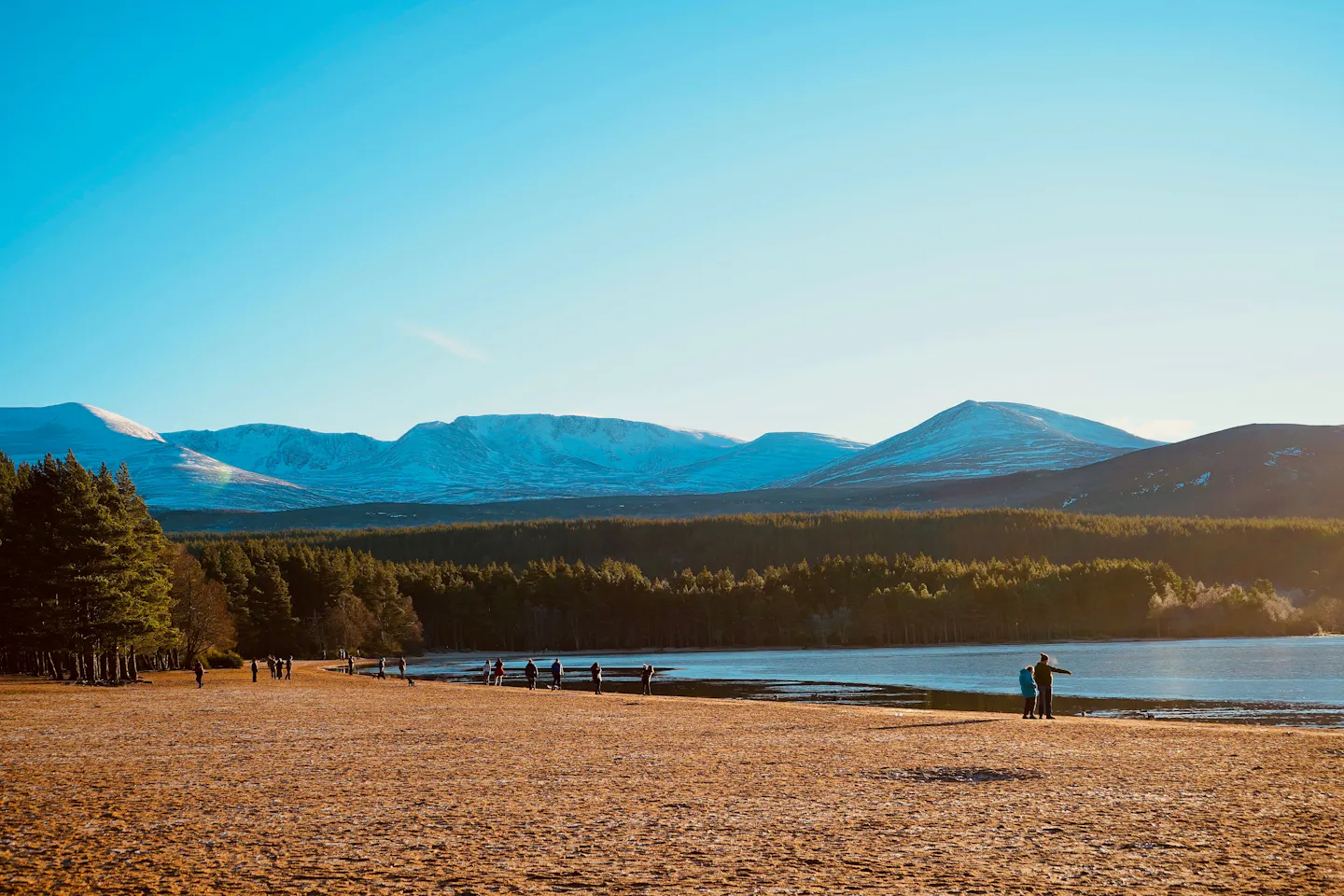 Walkers on the sandy shore of Loch Morlich below the snow-capped Cairngorms