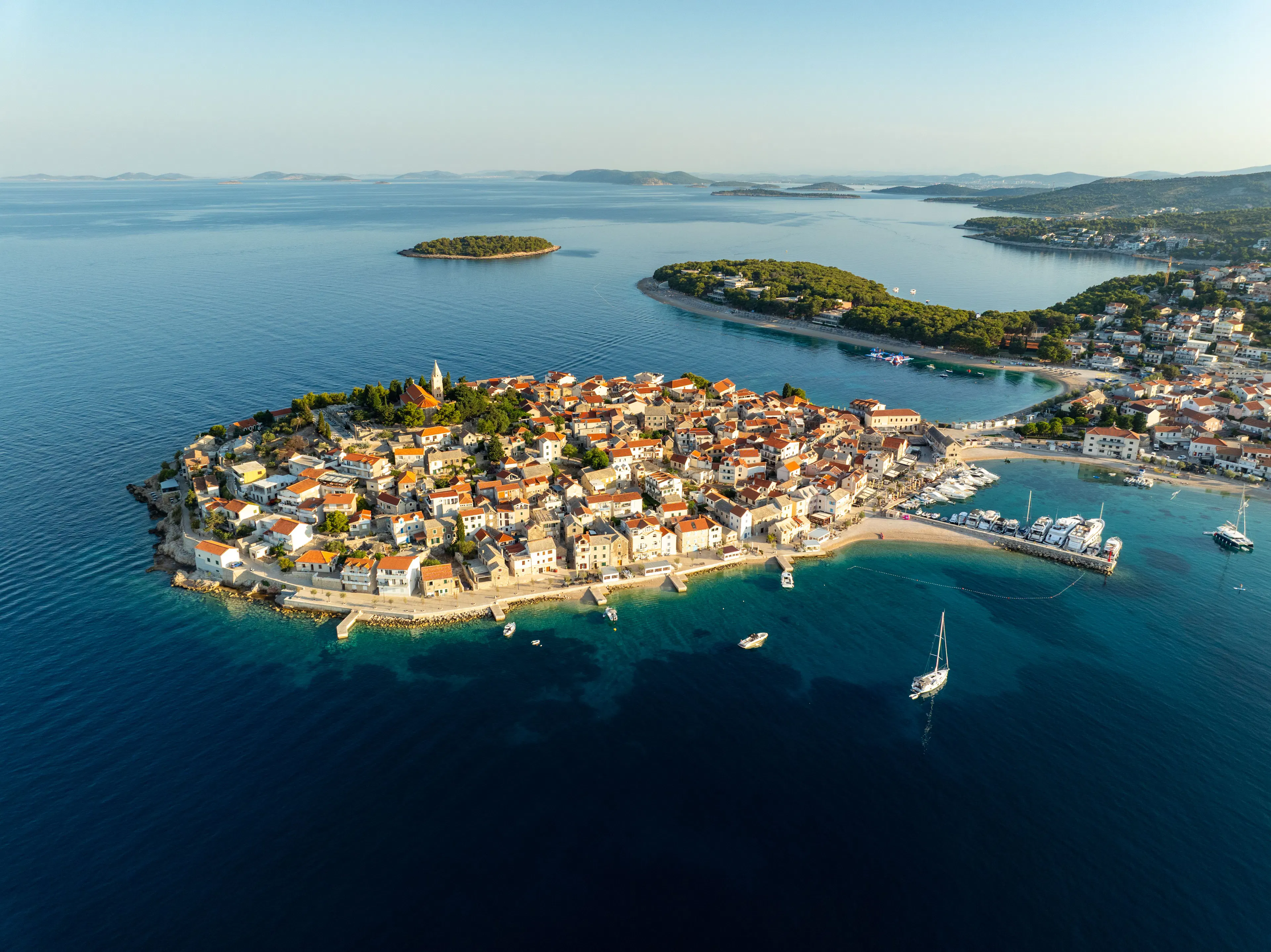 Aerial view of a red-roofed coastal town on a wooded peninsula, surrounded by blue Adriatic sea, small islands and boats