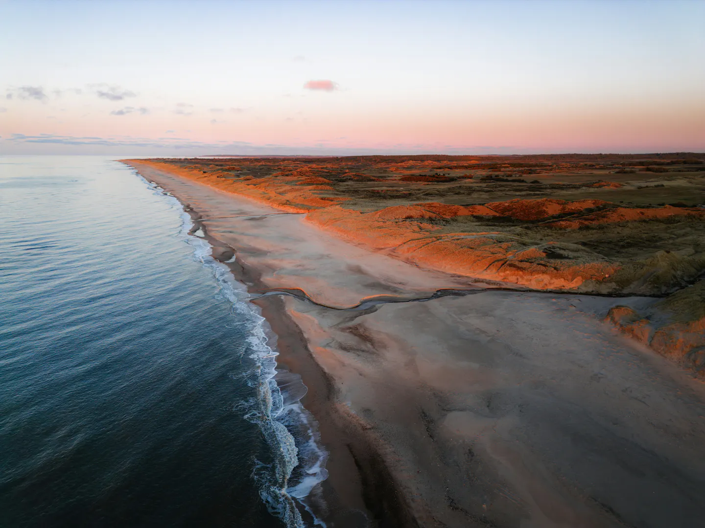 Long empty sandy beach curving beside grassy dunes under a pink and gold sunset sky