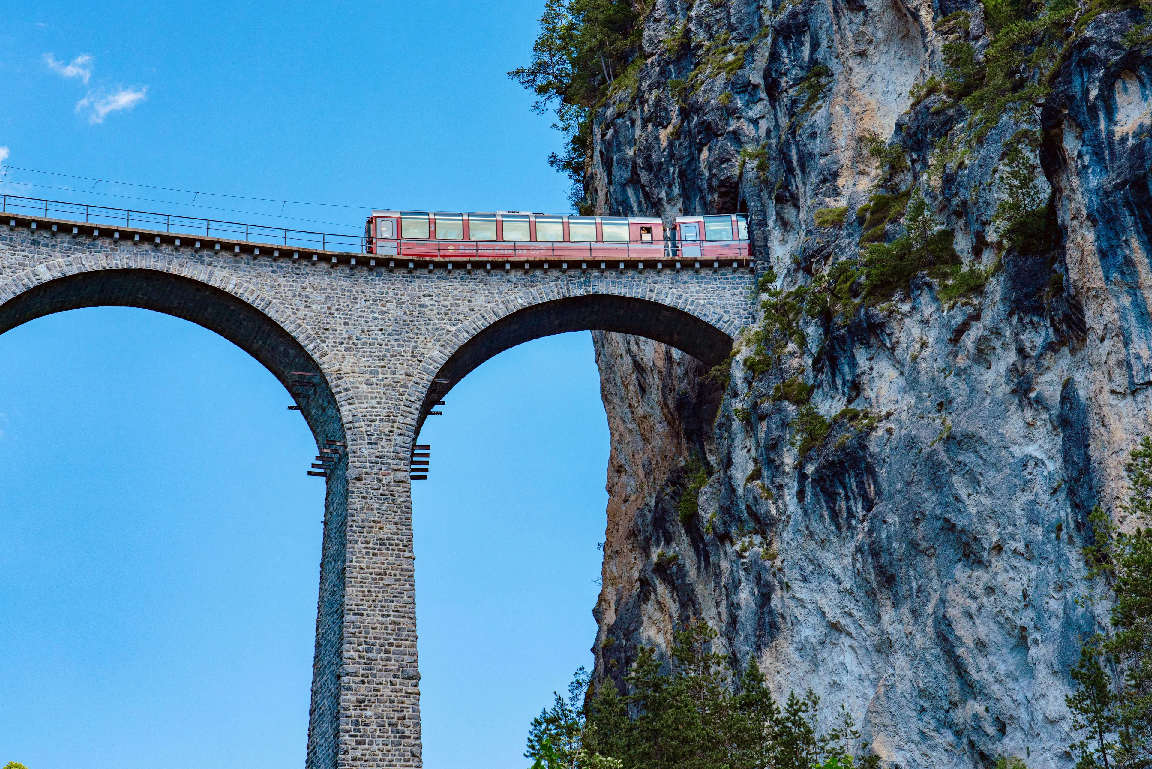 A red mountain train crossing a tall stone railway viaduct between rocky cliffs in the Swiss Alps