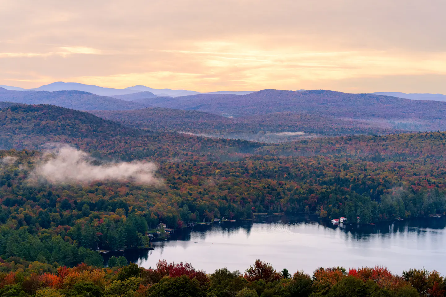 Mist over a lake ringed by forests in full autumn colour, with pale mountains at dawn