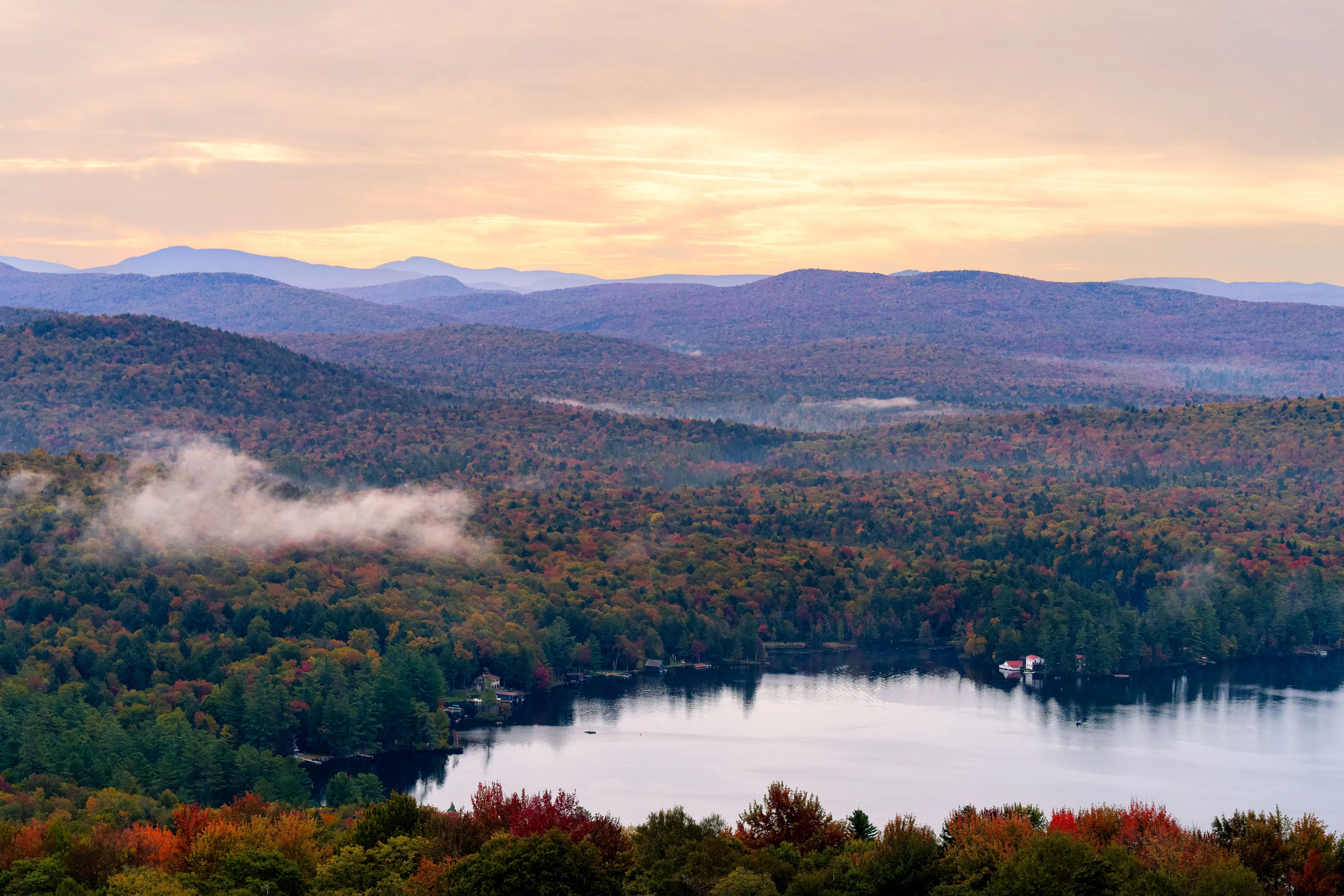 Mist over a lake ringed by forests in full autumn colour, with pale mountains at dawn