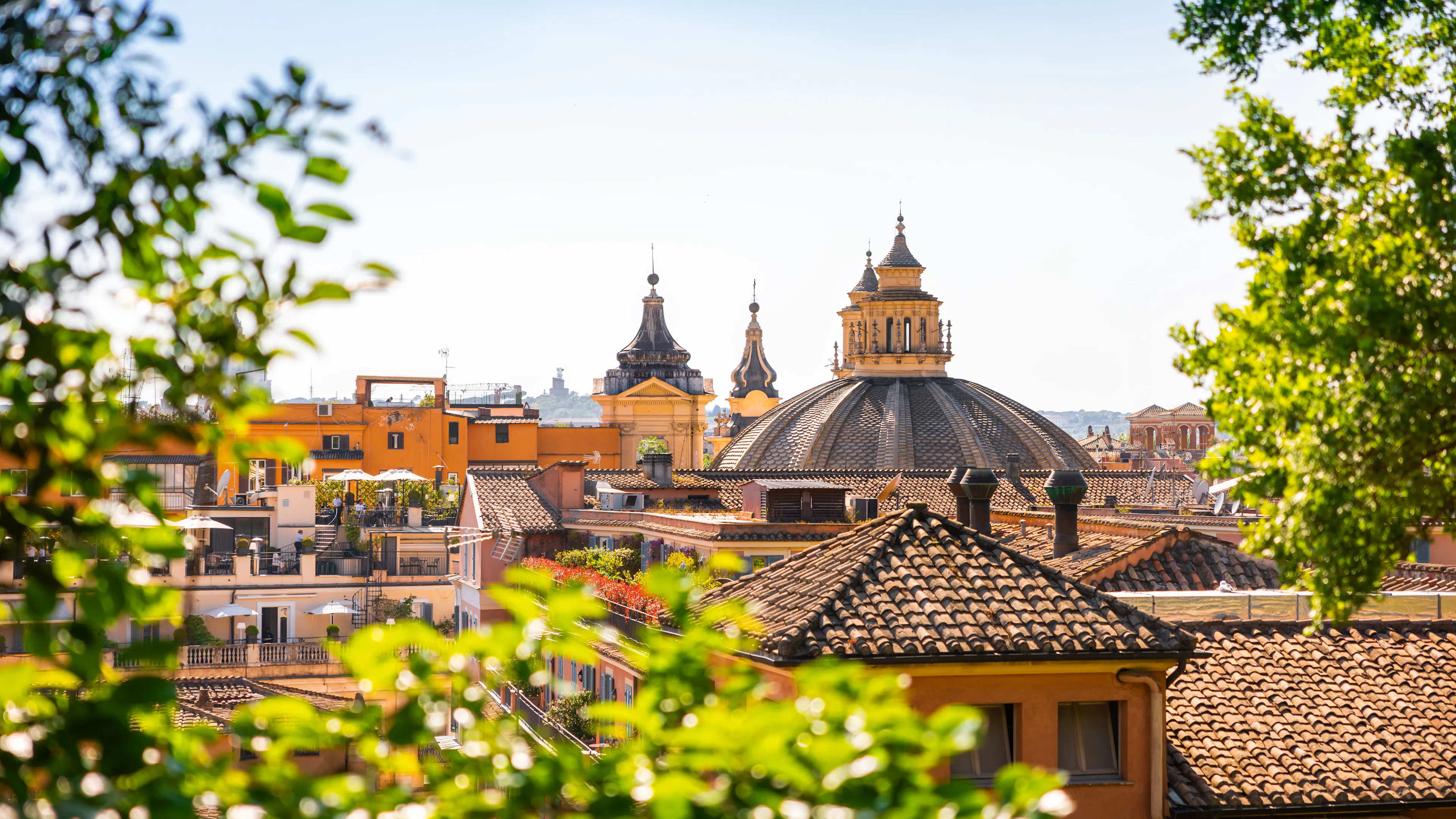 Terracotta rooftops, roof terraces and two baroque church domes in central Rome, framed by sunlit green leaves under a clear sky
