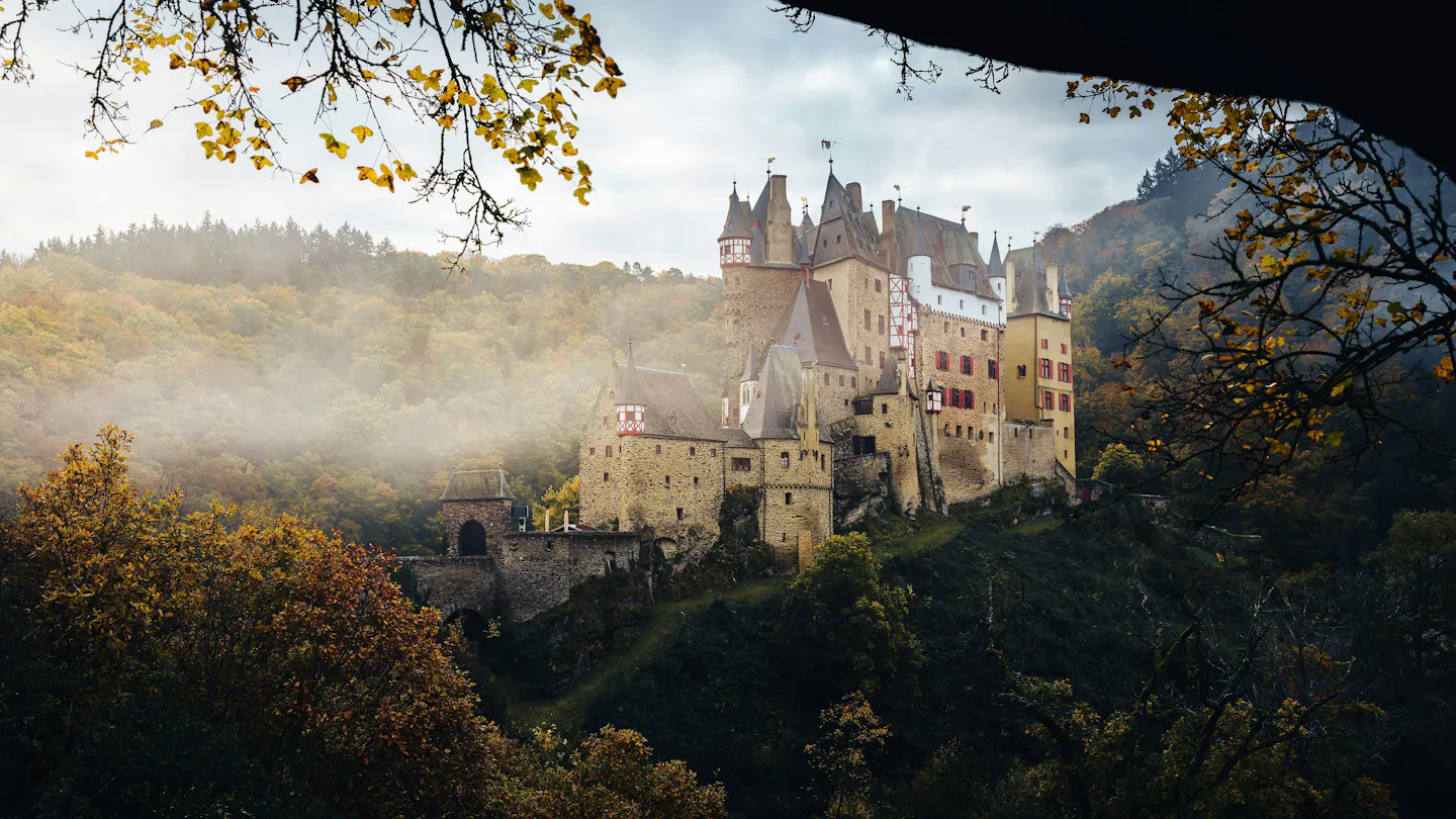 A many-turreted medieval castle standing on a wooded spur above a valley of autumn fog, framed by golden oak leaves and a dark rock overhang in the foreground