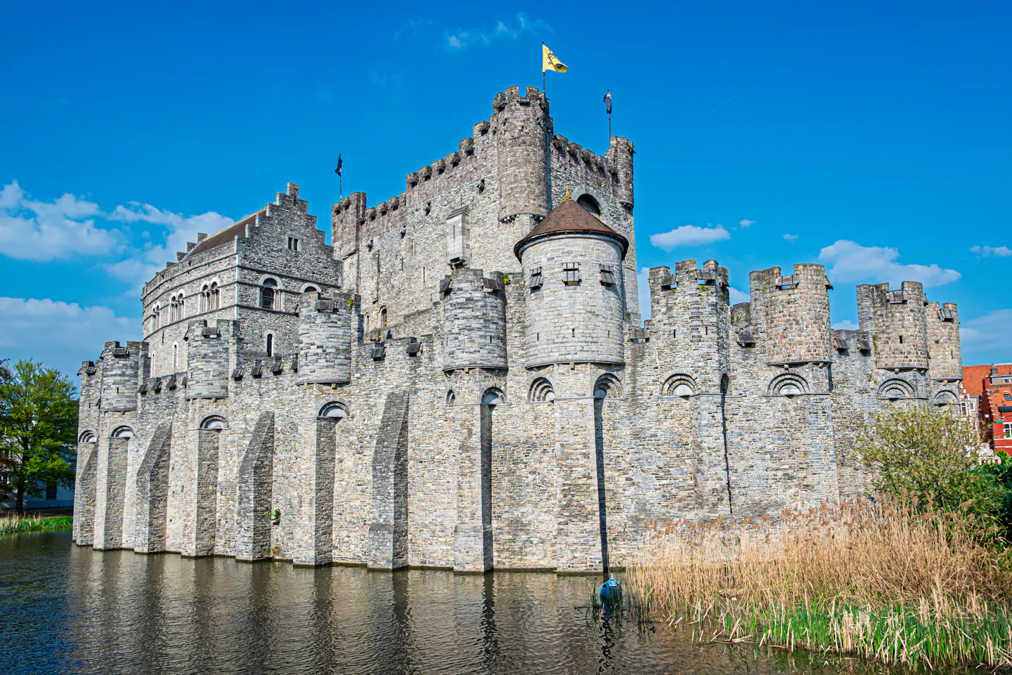 The stone walls and turrets of Gravensteen castle reflected in its moat in Ghent under a blue sky