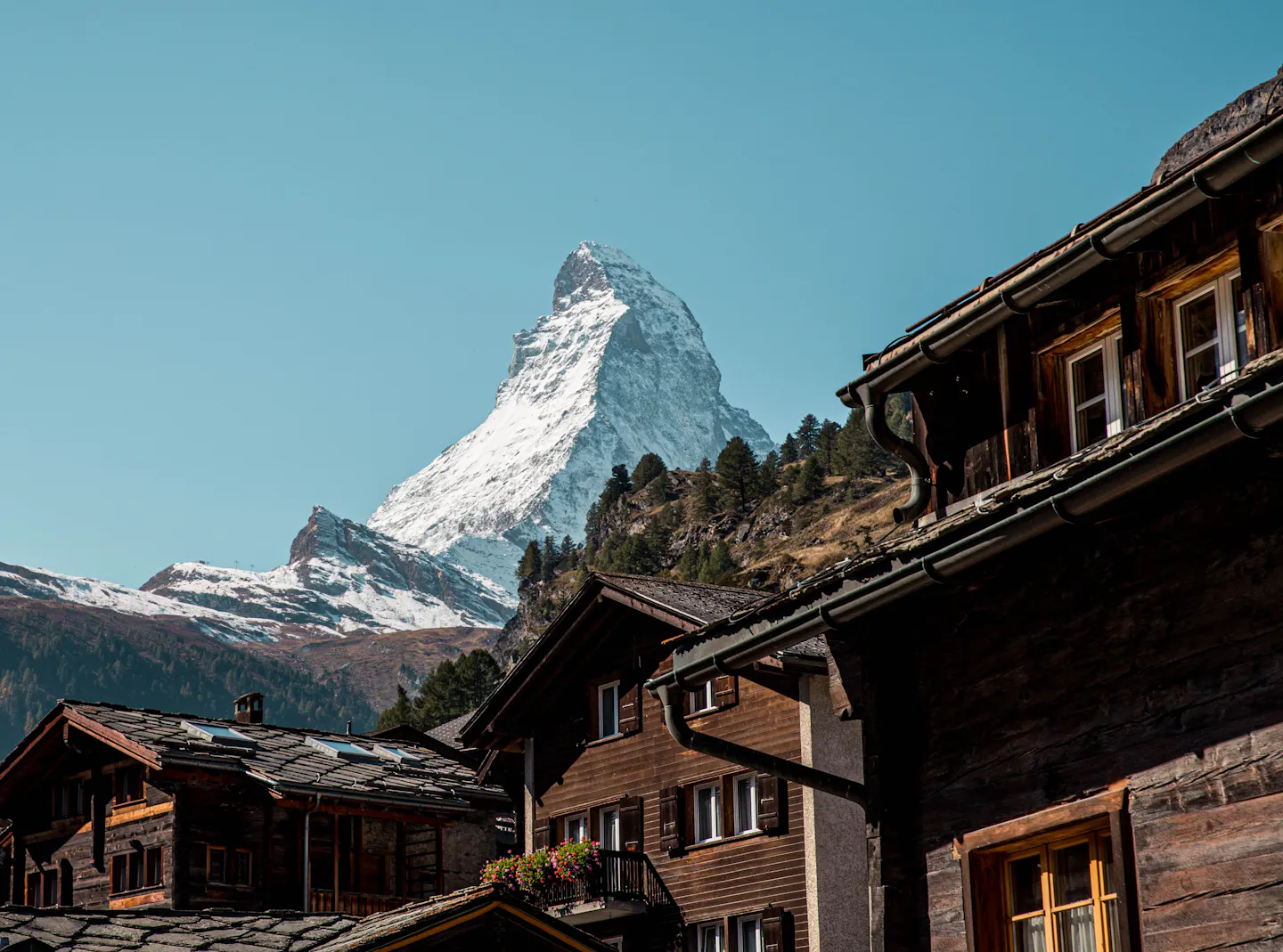 Dark-timber Zermatt chalets with flower boxes and slate roofs in the foreground, the snow-covered Matterhorn rising directly behind under a deep blue sky