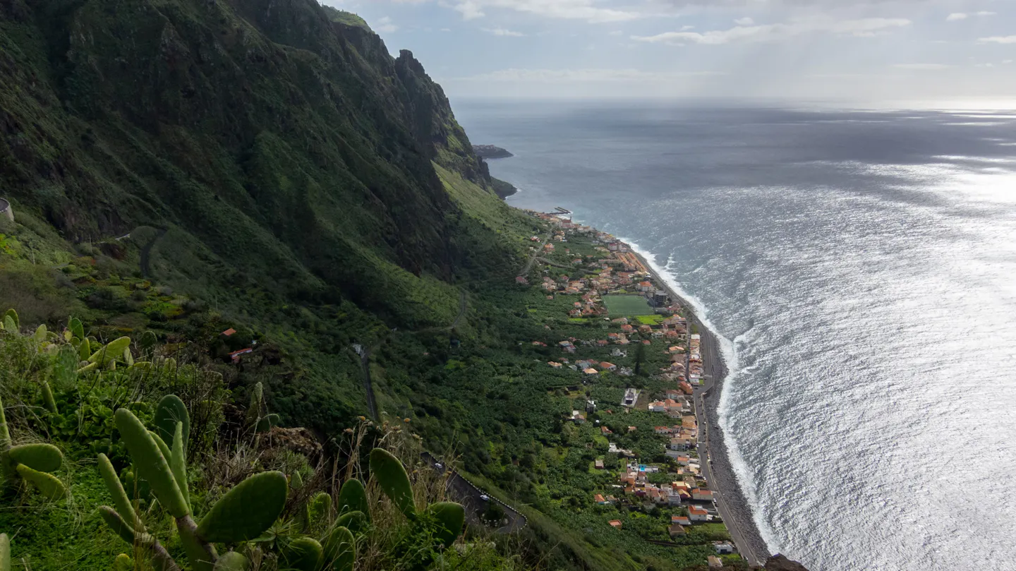 A terraced village strung along Madeira’s shoreline beneath towering green volcanic cliffs, the Atlantic breaking white along the shore